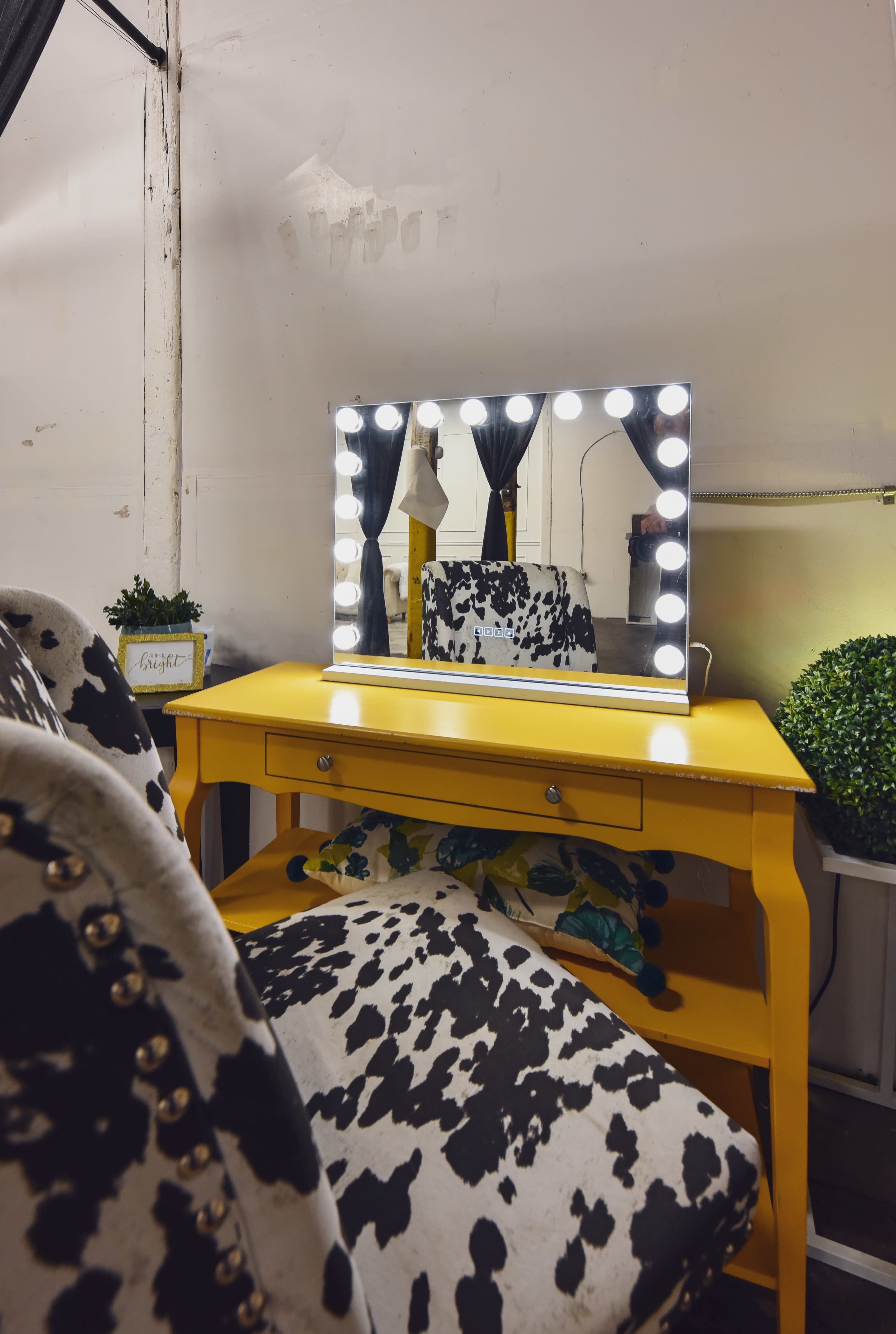 The image shows a yellow table with a cow-print chair and a lit makeup mirror set against a plain wall.