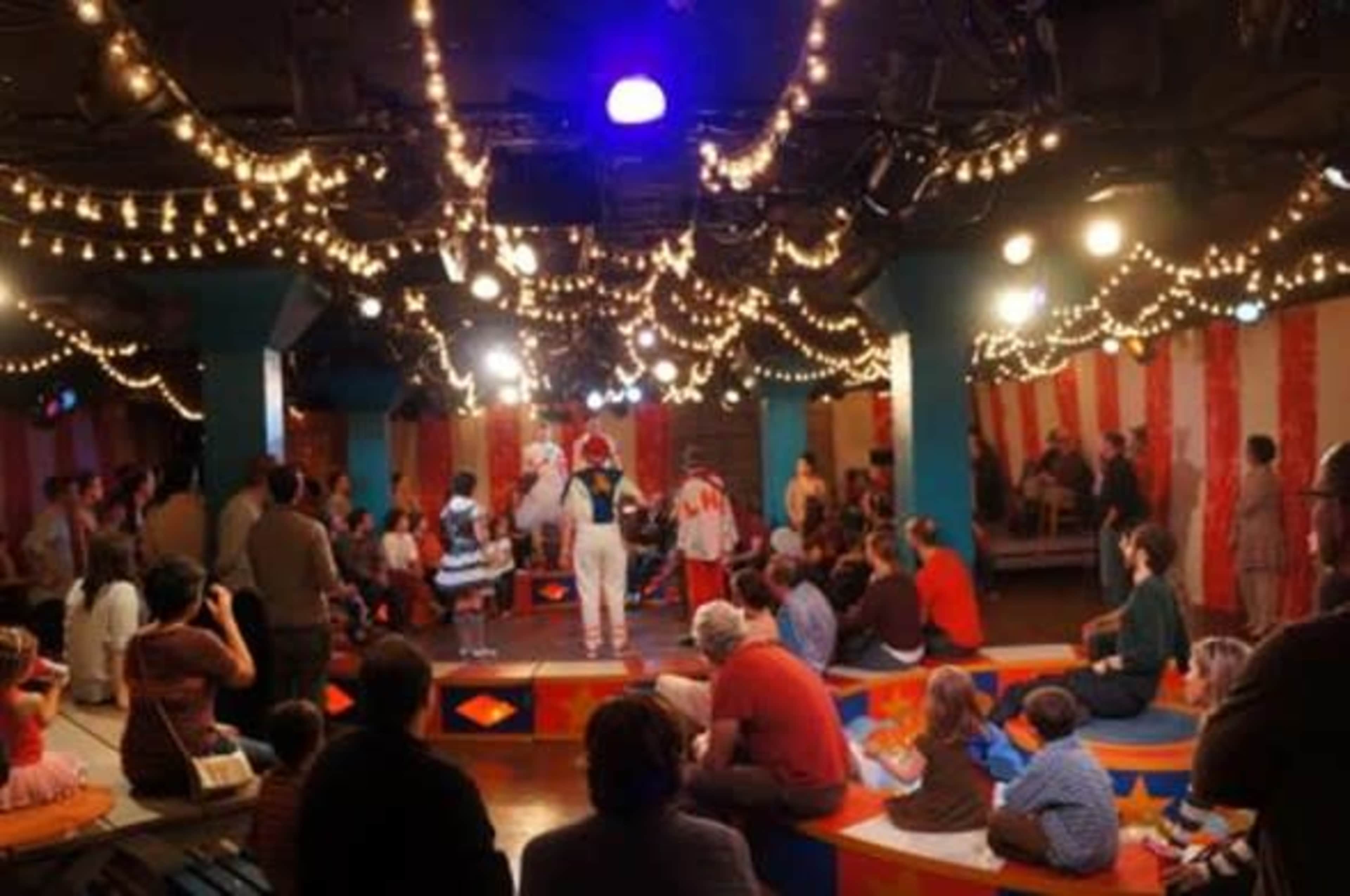 A vibrant circus-themed performance is taking place indoors, with performers in costumes entertaining a diverse audience seated on colorful platforms under string lights.
