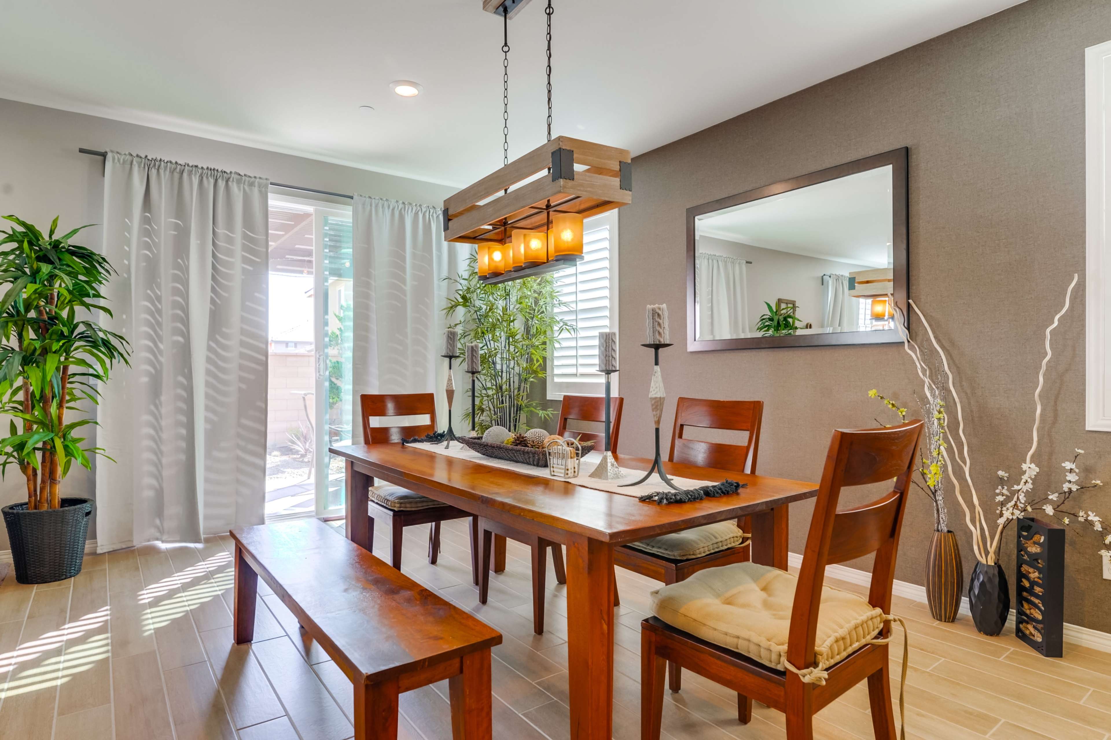 A dining room features a wooden table surrounded by chairs and a bench, with a mirror and plants against the wall.
