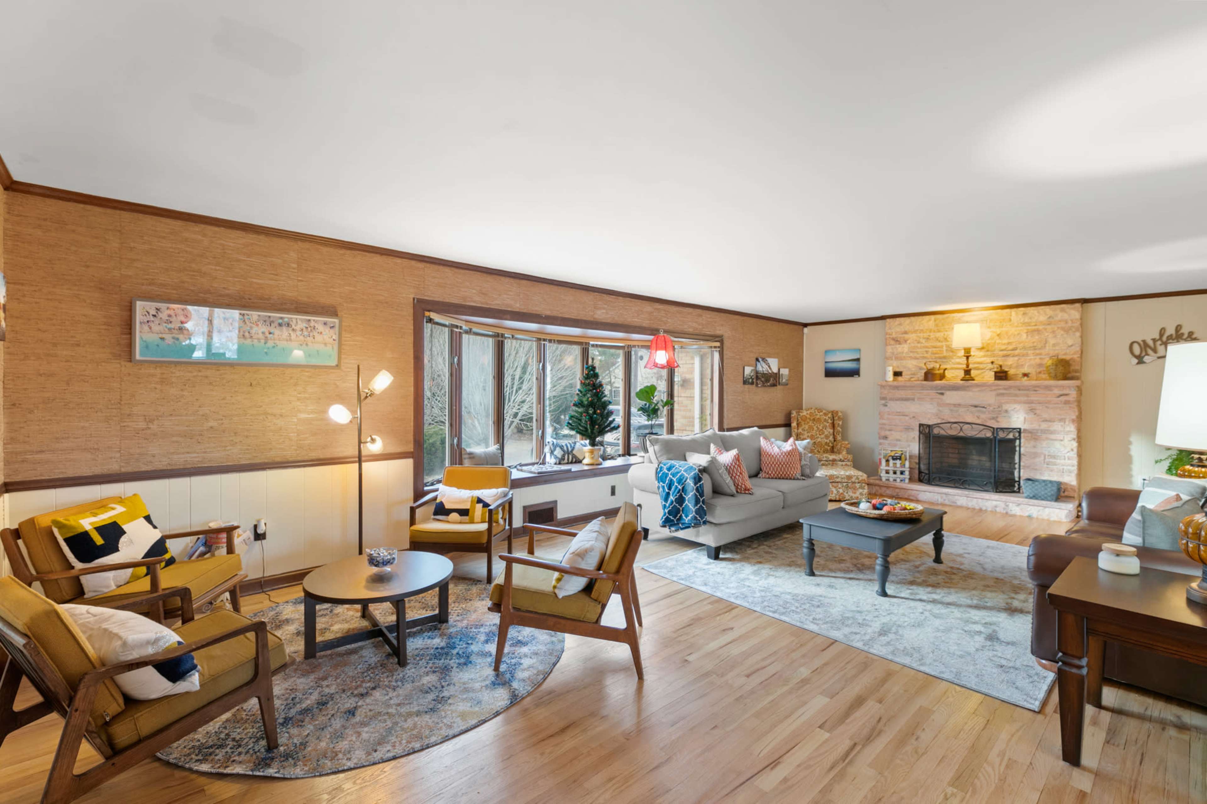 A well-lit living room features a cozy seating area with a sofa and armchairs, a coffee table, and a stone fireplace, surrounded by large windows.
