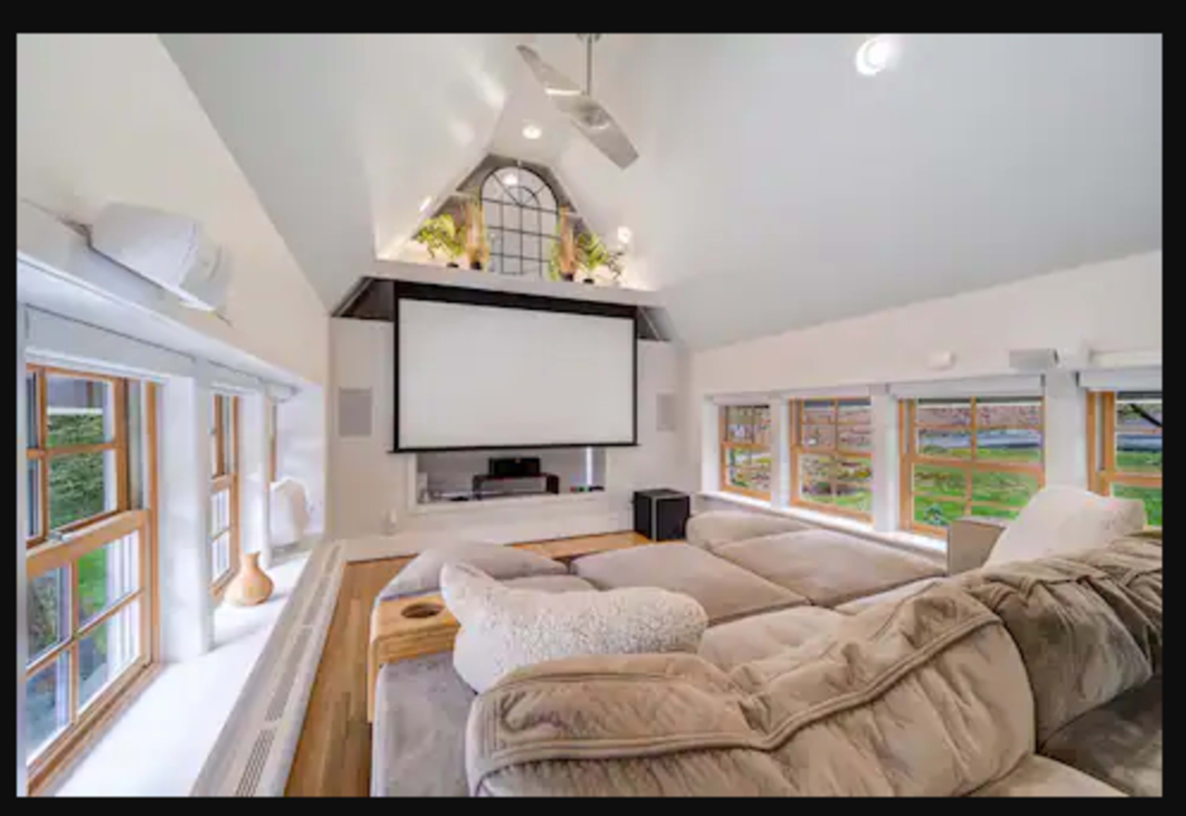 The room features a large sectional sofa facing a wall-mounted projector screen, with windows providing natural light and a view of the outdoors.