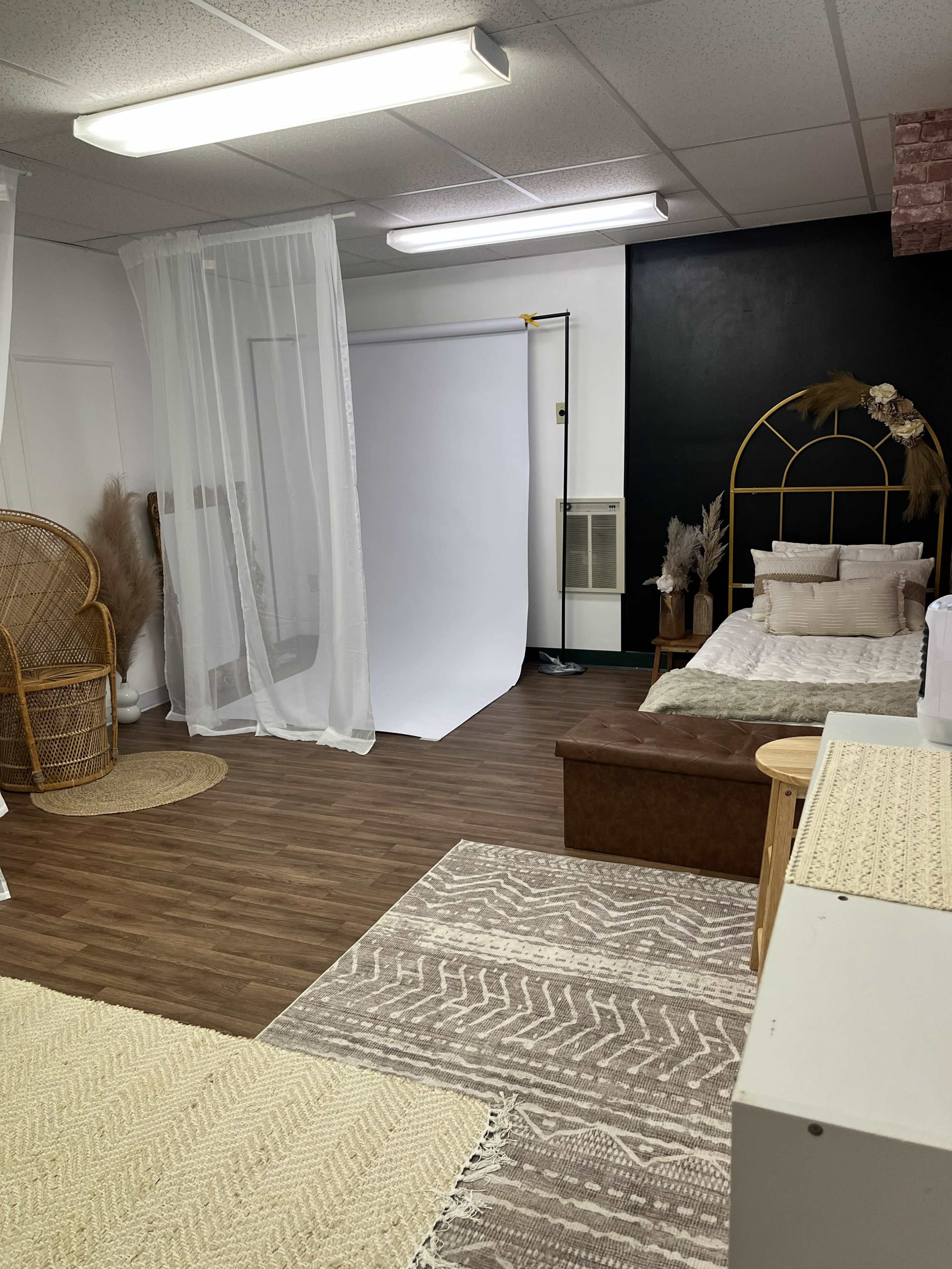 The image shows a styled indoor space featuring a bed, a wicker chair, various rugs, and a backdrop for photography.