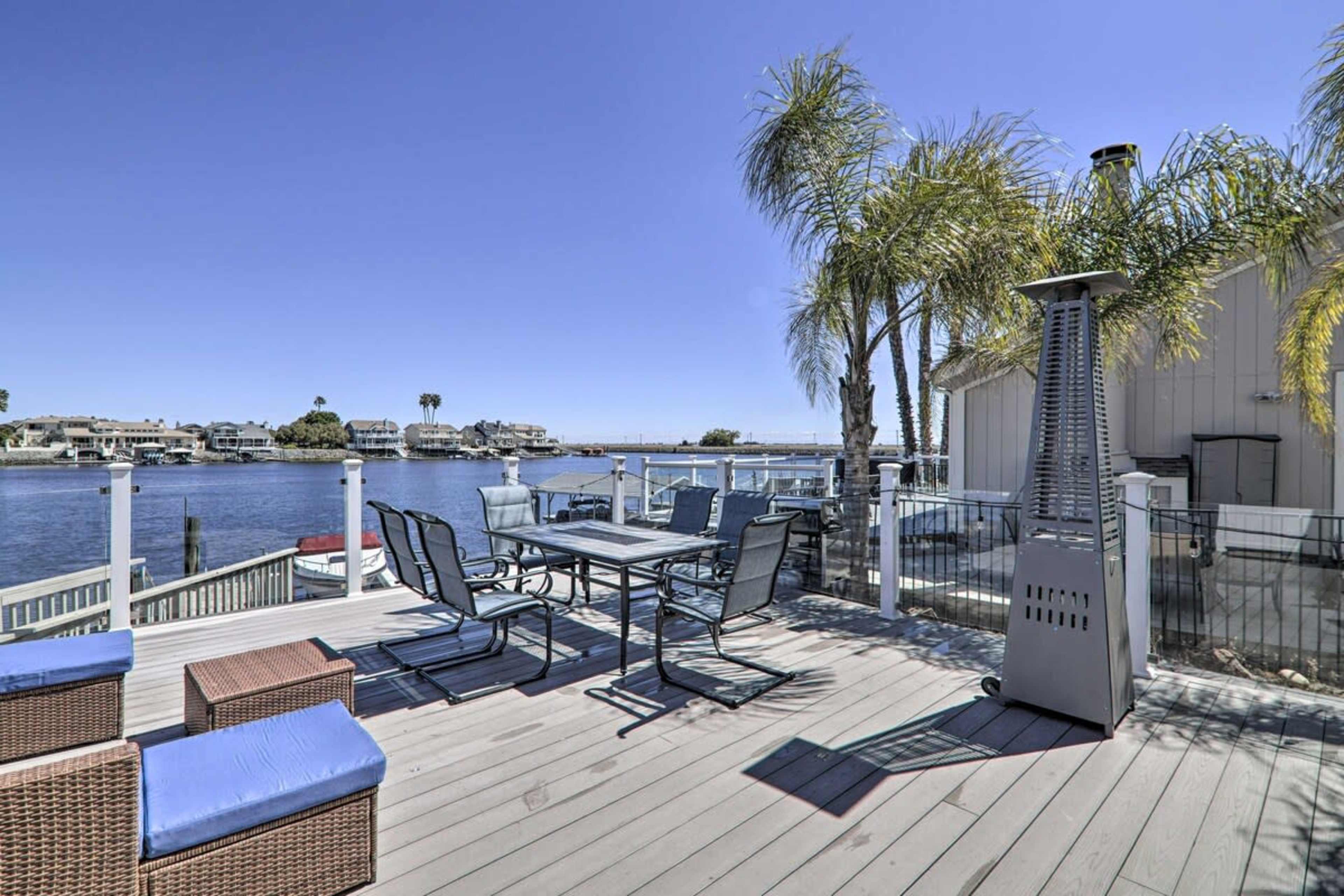 A wooden deck overlooks a calm waterfront with palm trees and dining furniture.