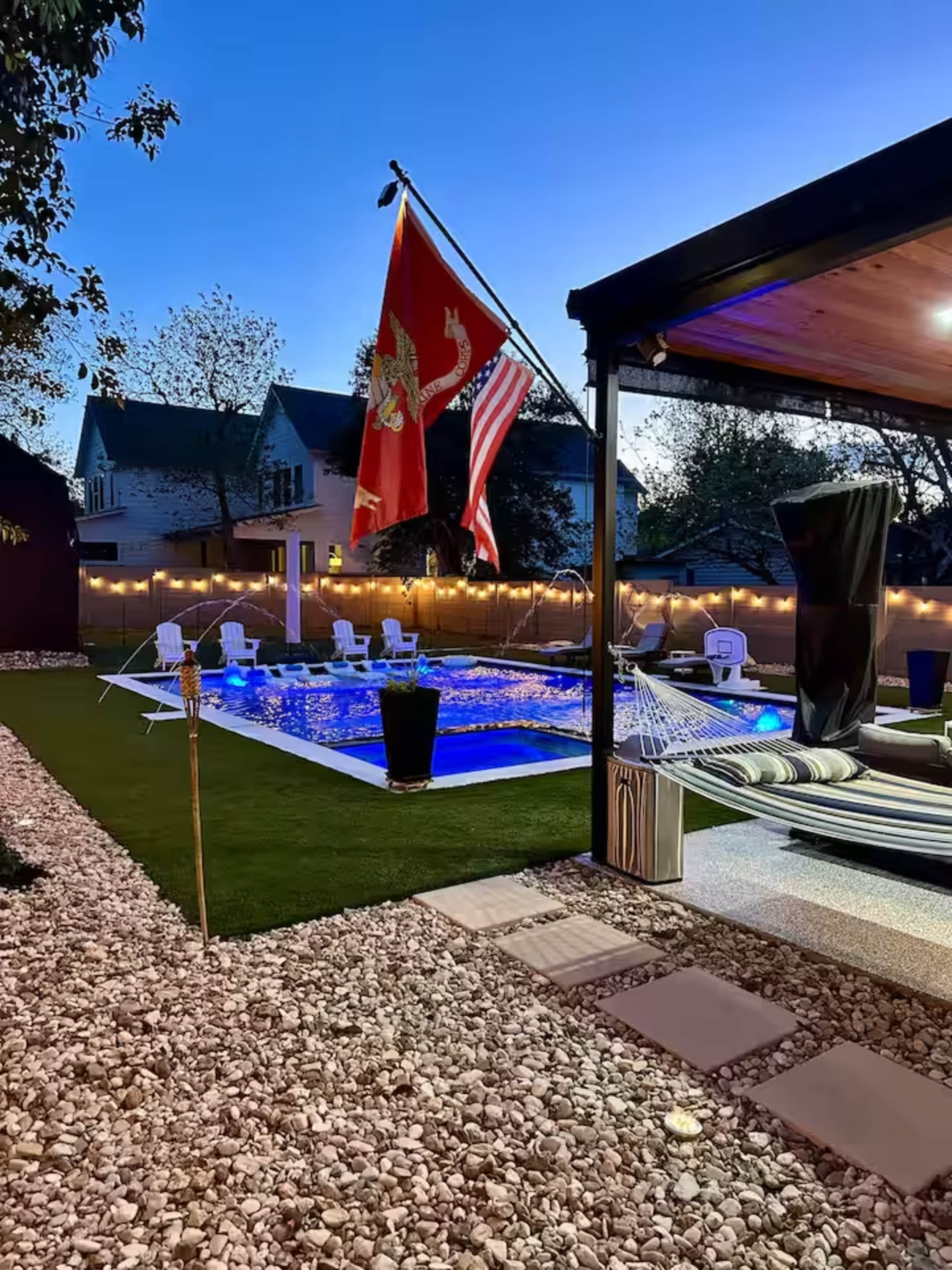 The image shows a backyard pool area with illuminated water, flags flying, and modern seating arrangements surrounded by decorative rocks and landscaping.