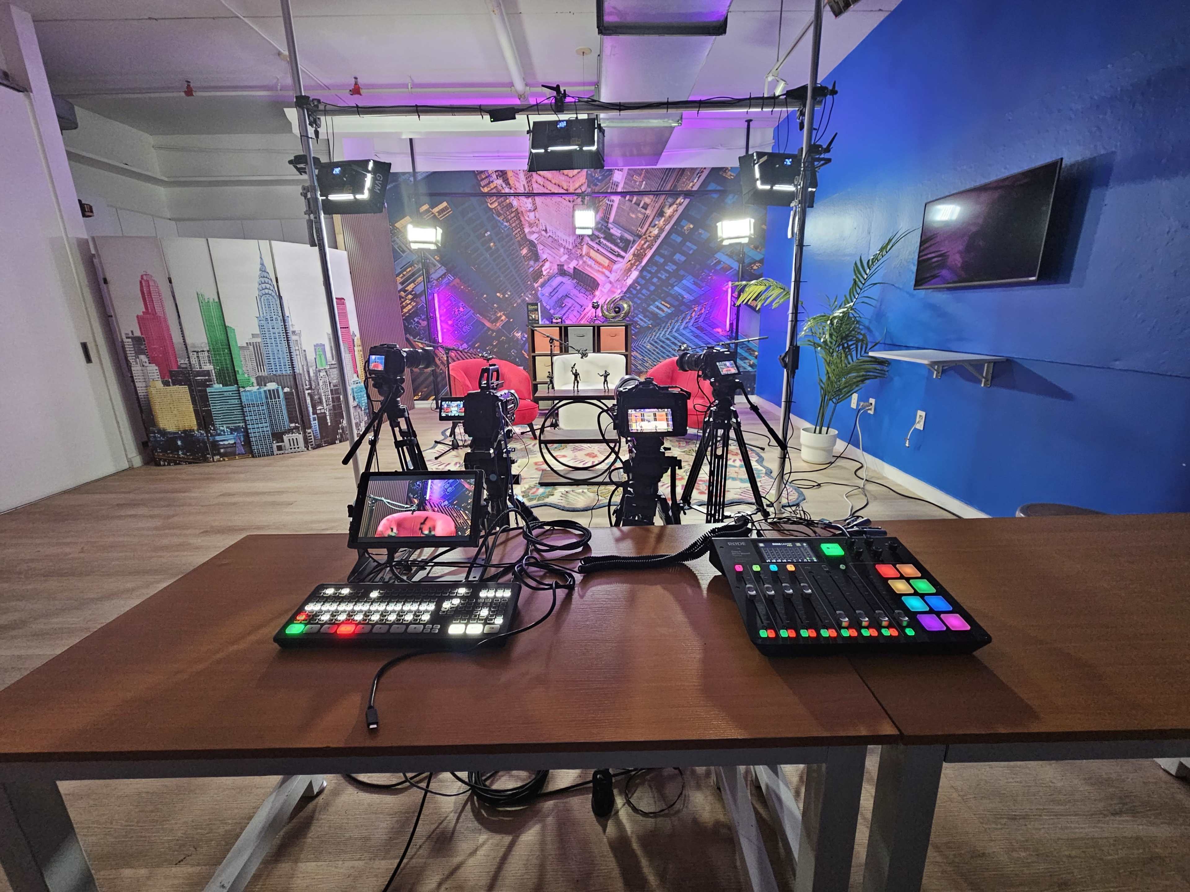 The image shows a video production studio equipped with multiple cameras, lighting setups, and a control table featuring various audio-visual controls.