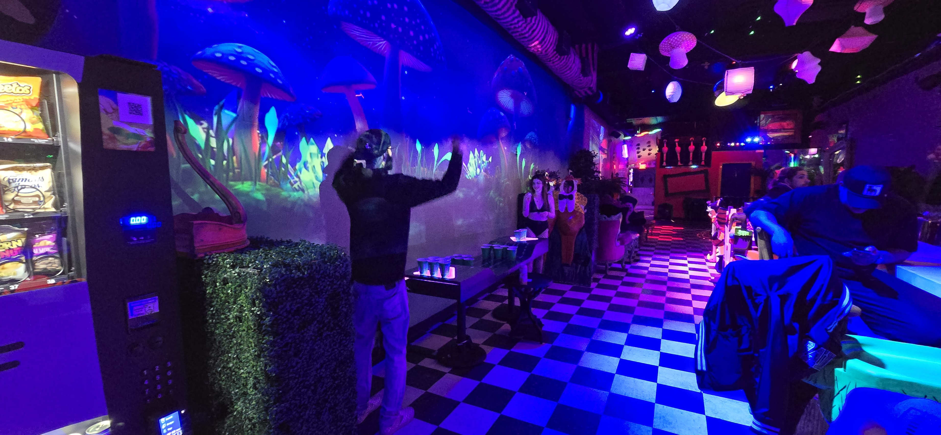 A person with a virtual reality headset stands in a colorful, dimly lit lounge decorated with mushroom murals and a checkered floor.