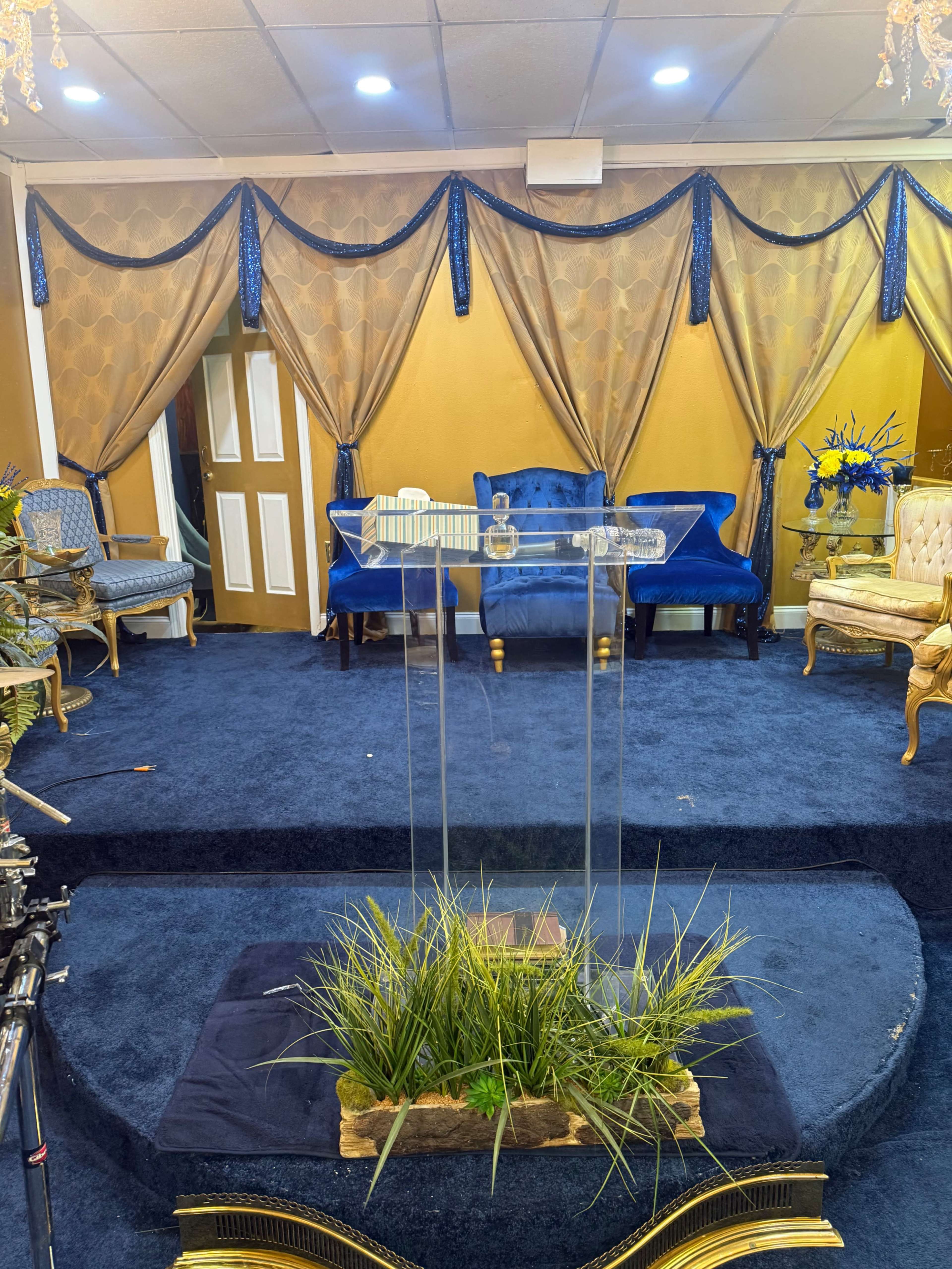 The image shows an indoor space designed for a gathering, featuring a clear podium at the center, surrounded by blue and gold decorations, with seating arrangements in the background.