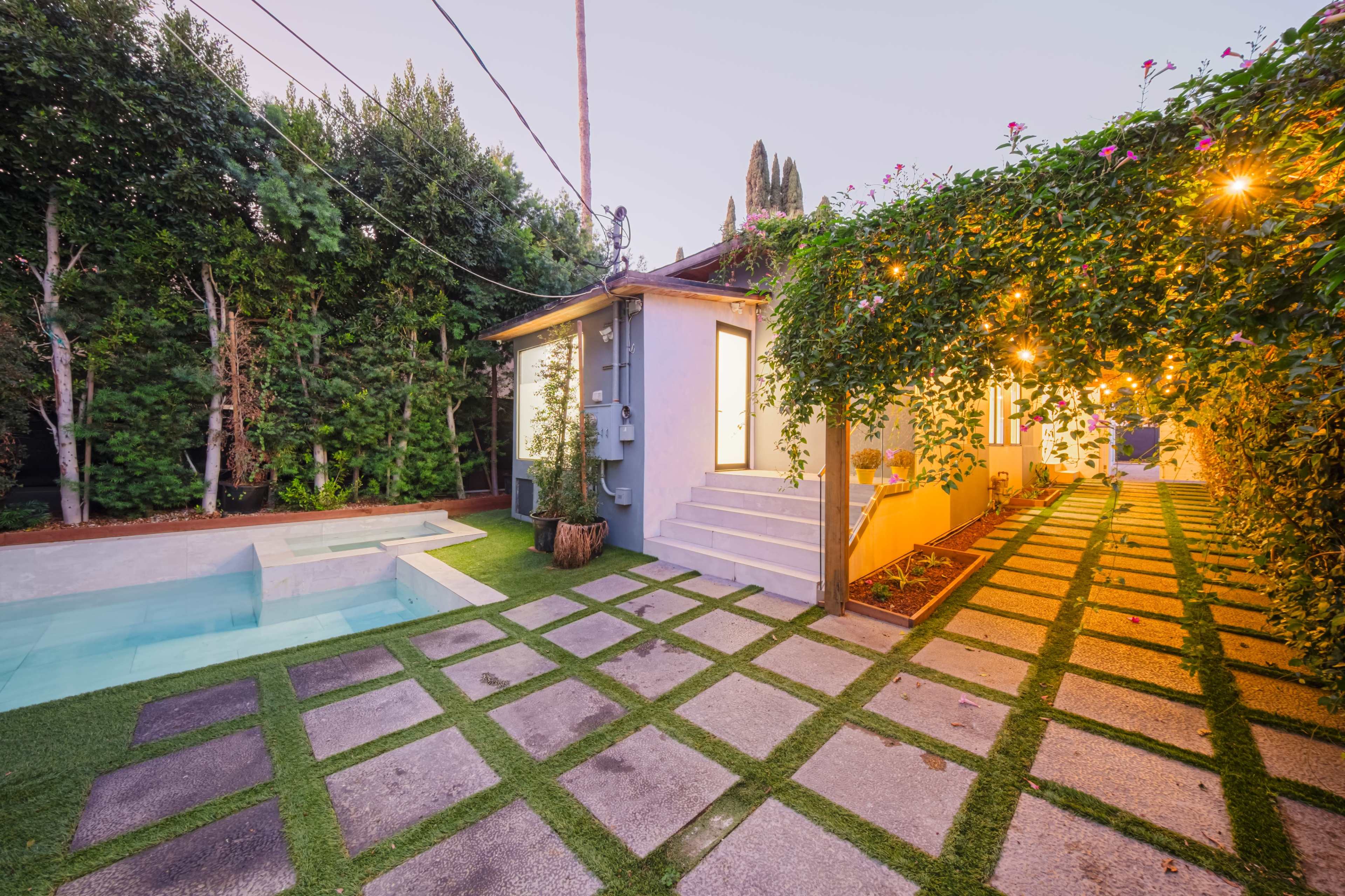 The image shows a backyard scene featuring a small pool, a stone pathway with square grass patches, and a modern, well-lit building surrounded by lush greenery.