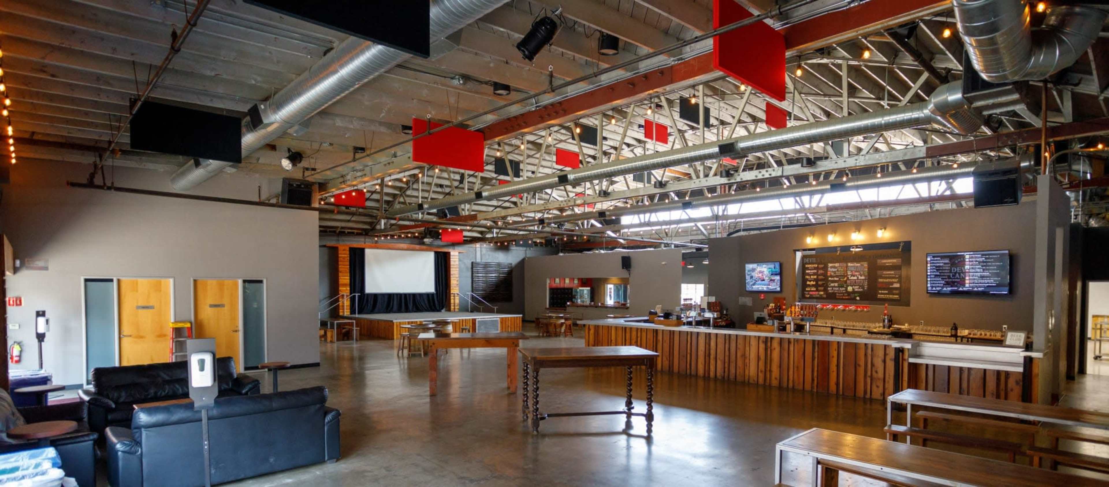A spacious, modern interior of a venue with high ceilings, wooden tables, and a bar area featuring screens and a menu board.