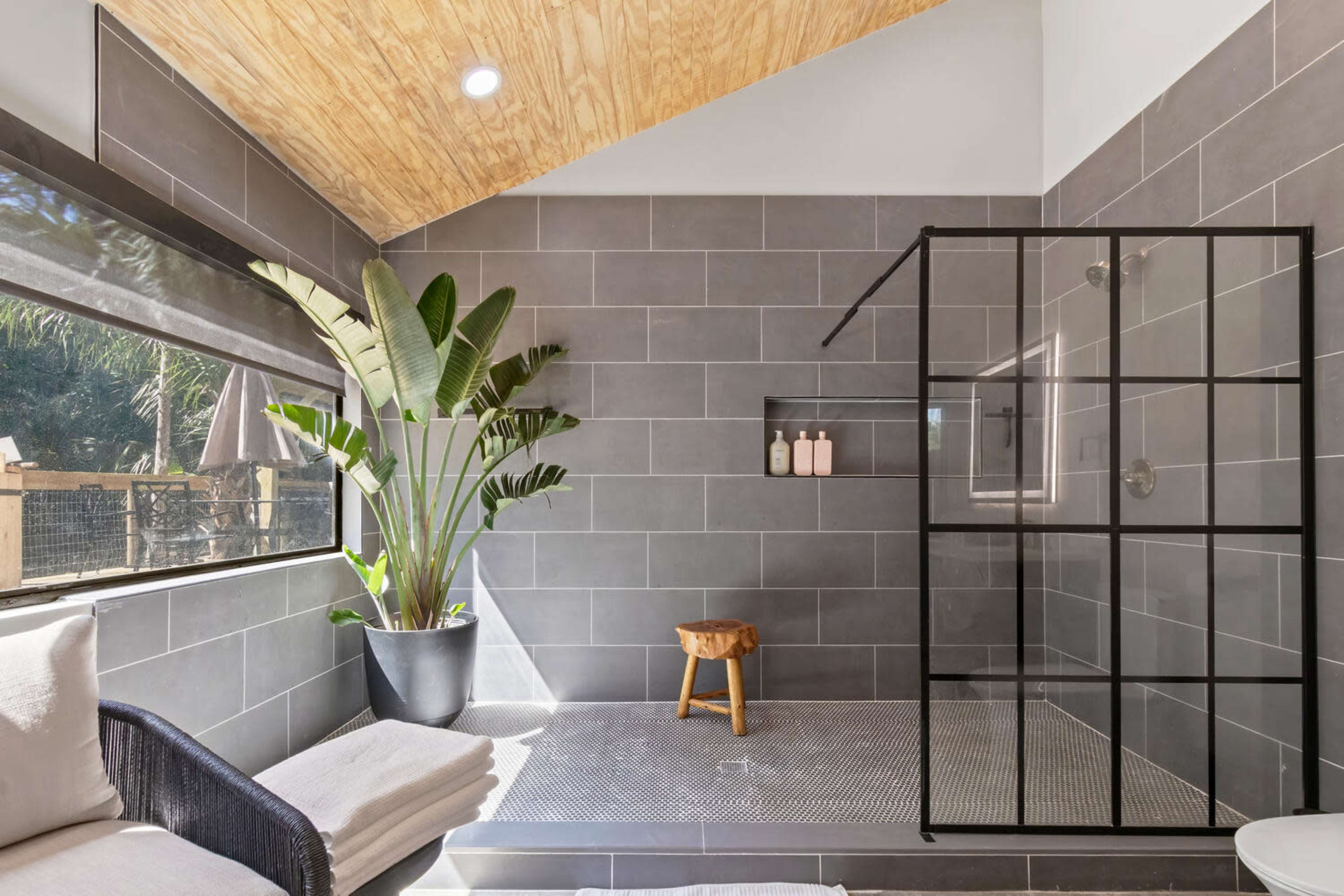 The image shows a modern bathroom featuring a glass shower enclosure, a wooden stool, and indoor plants, all set against gray tiled walls.