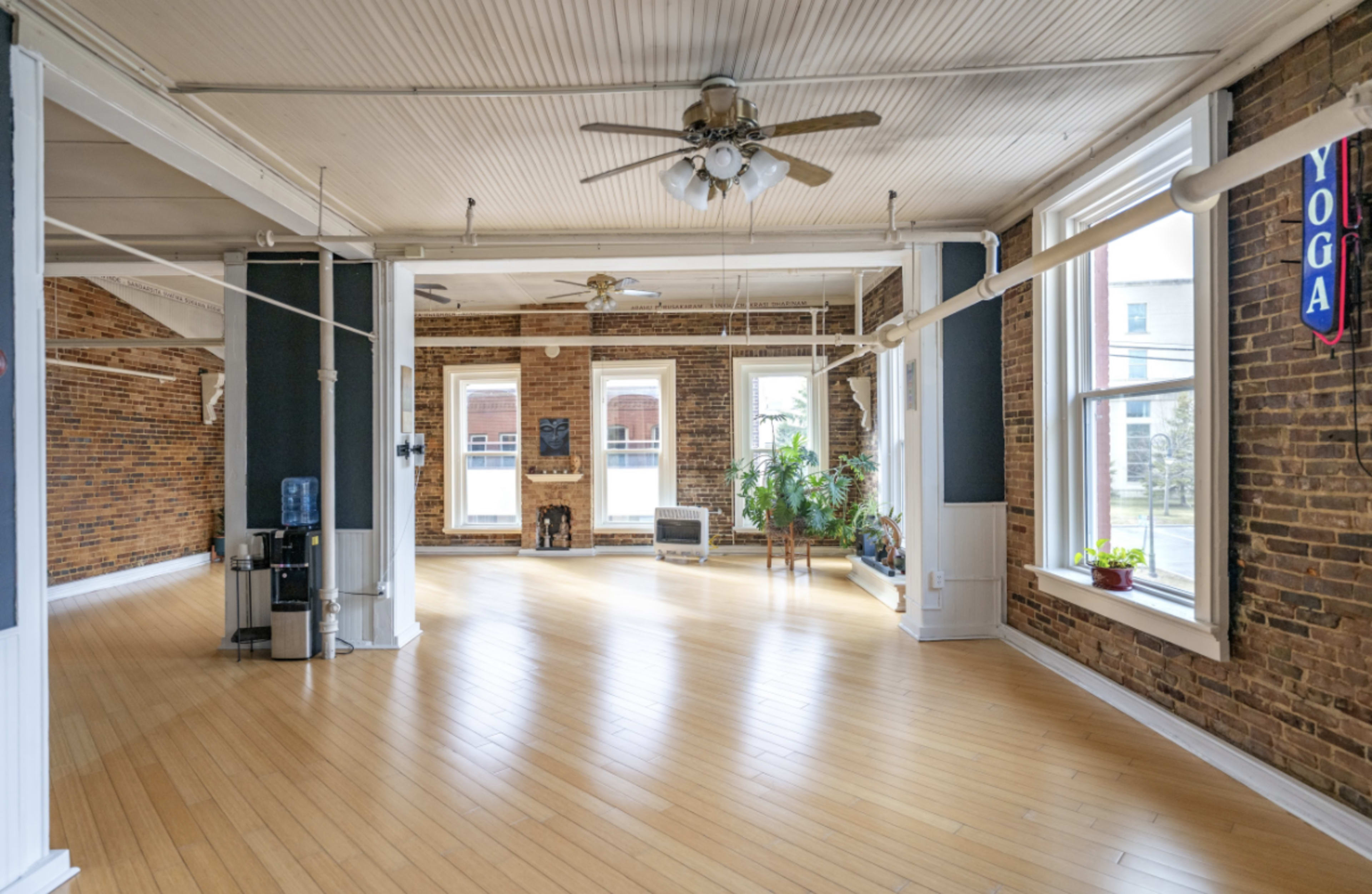 The image shows a spacious yoga studio with wooden floors, large windows, and exposed brick walls, featuring a water cooler and indoor plants.