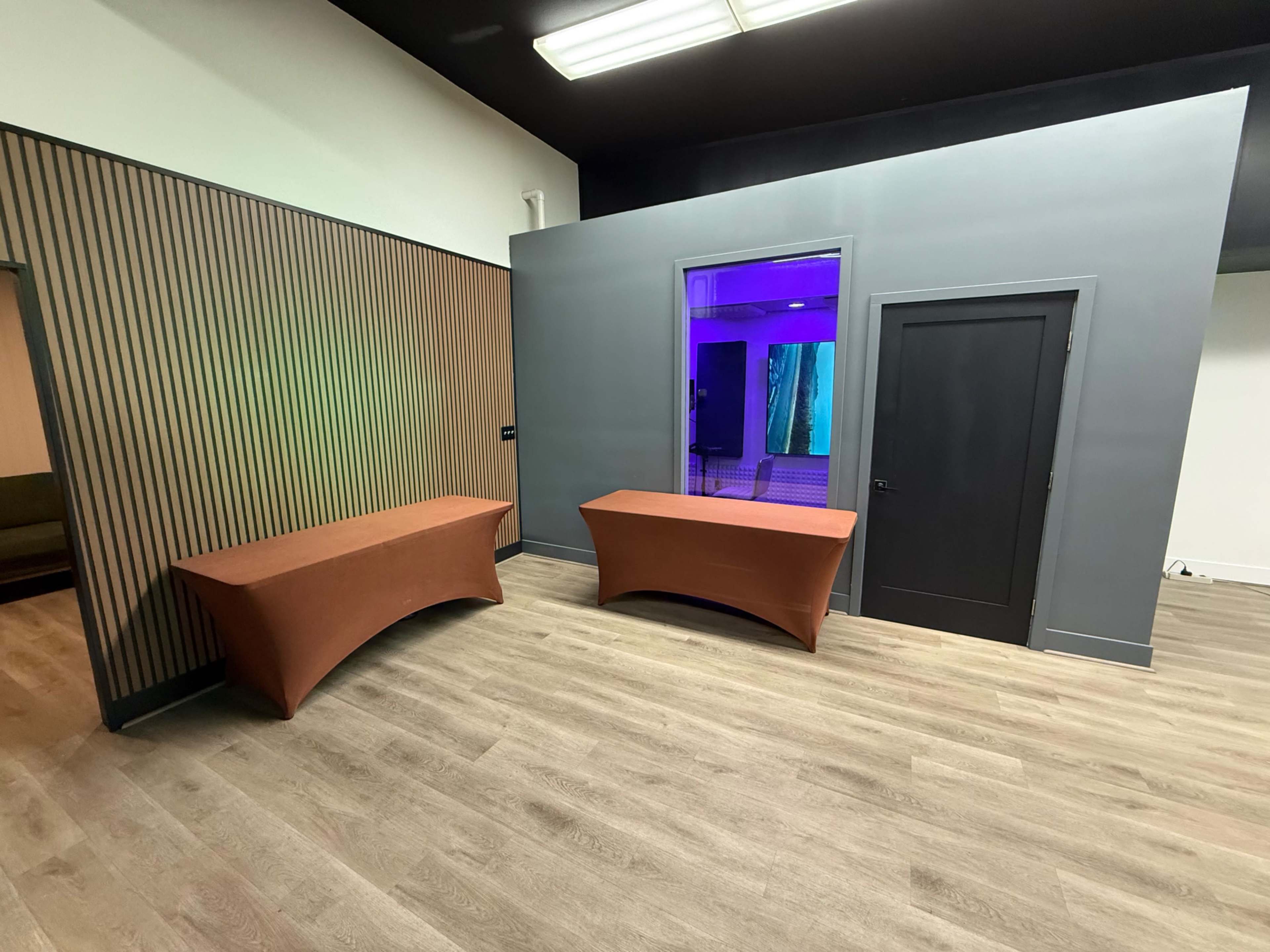 The image shows a modern interior space with two brown tables against a gray wall, and a doorway leading to a brightly lit area with purple and blue lighting.