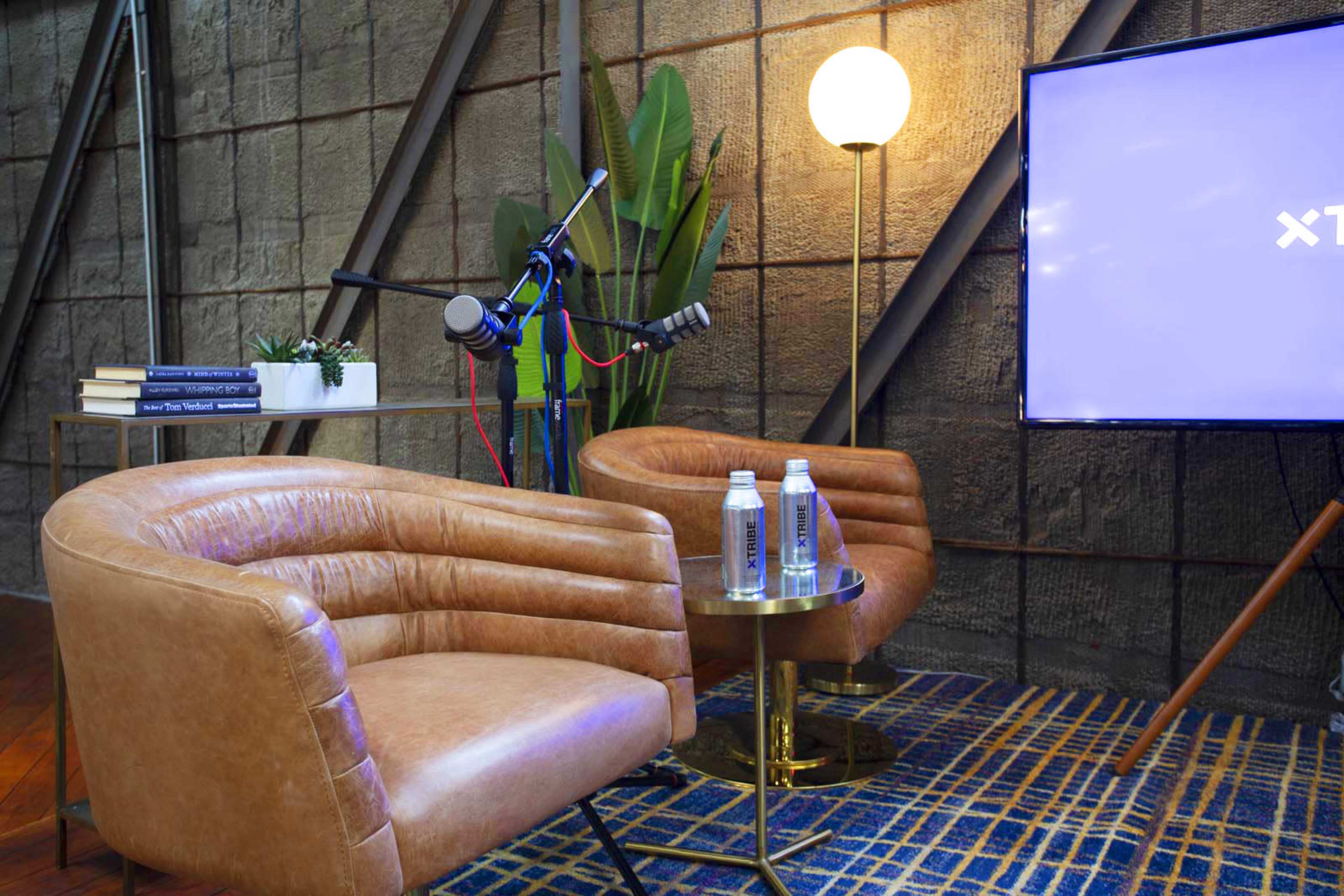 The scene features two brown chairs beside a small table with water bottles, a microphone on a stand, and a television screen in a modern interior setting.