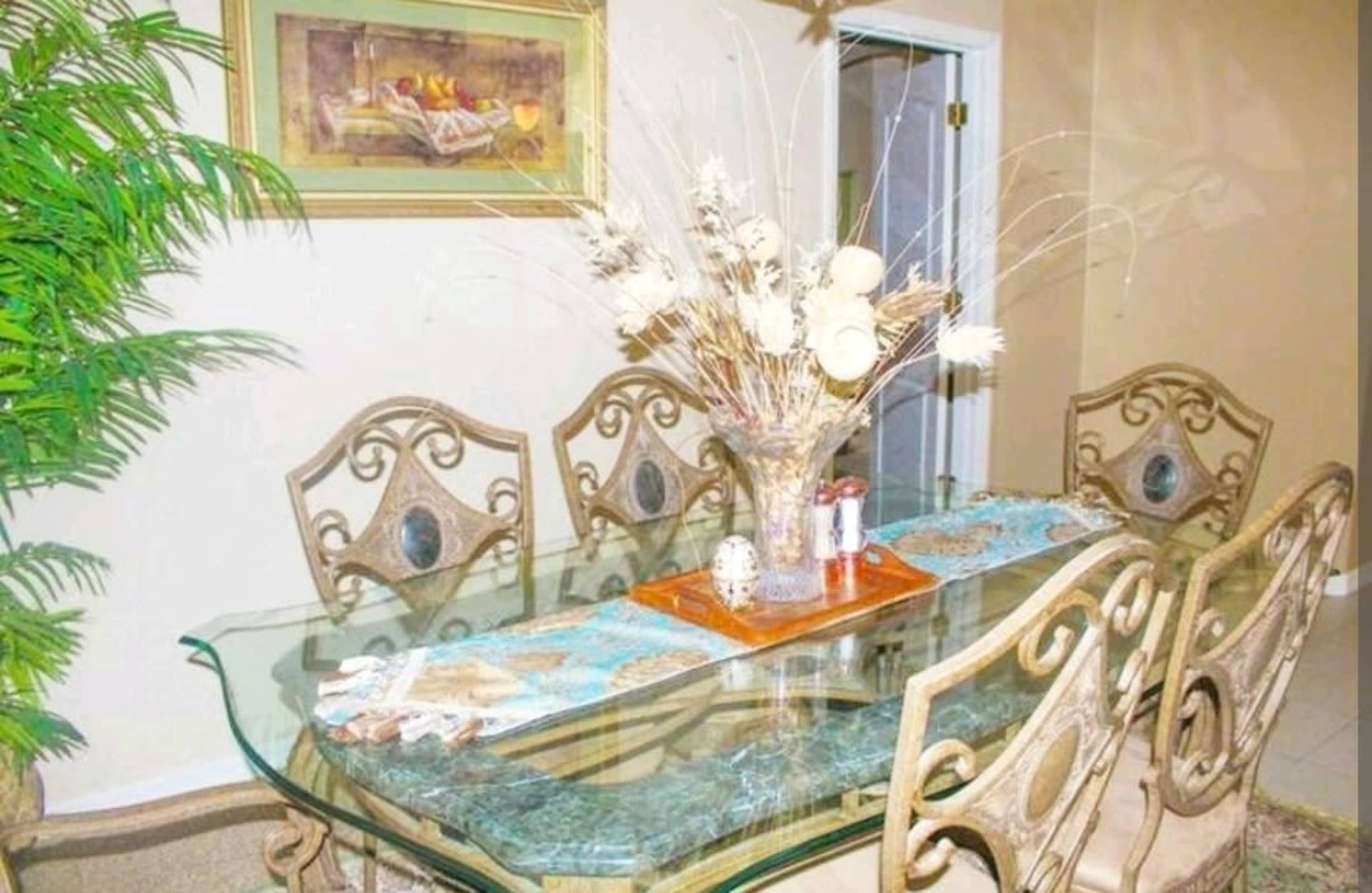 A glass dining table is set with a decorative centerpiece and surrounded by ornate metal chairs in a well-lit room.