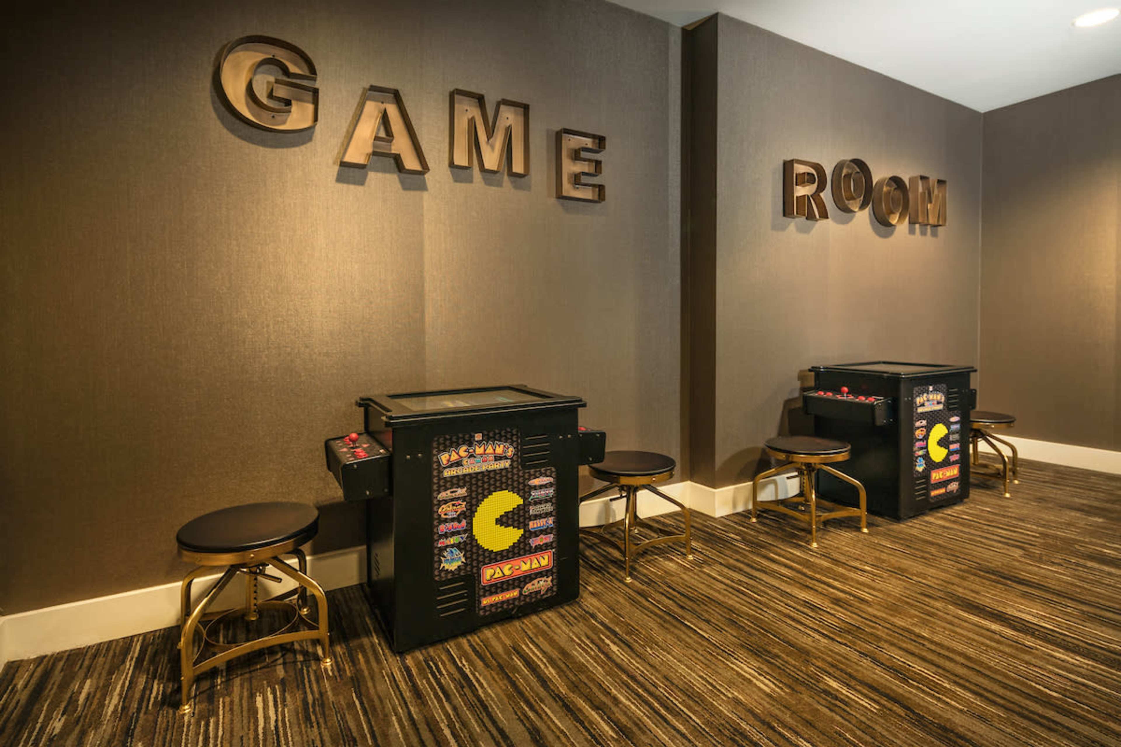 A game room with two arcade machines and seating arranged along a wall featuring large letters spelling "GAME ROOM."