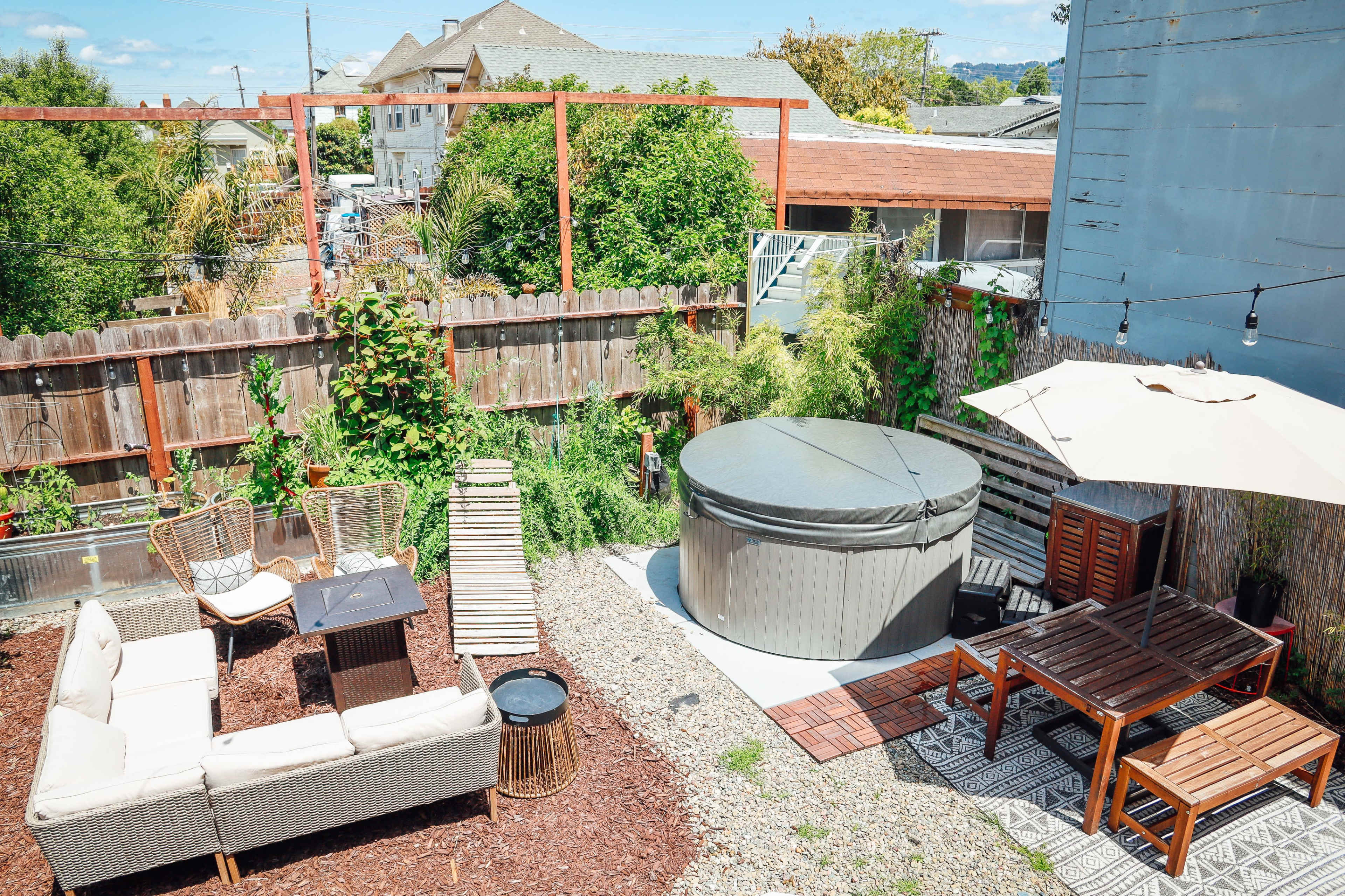 A backyard space featuring a hot tub, a wooden dining table, a seating area with a sofa, and landscaped greenery surrounded by wooden fencing.