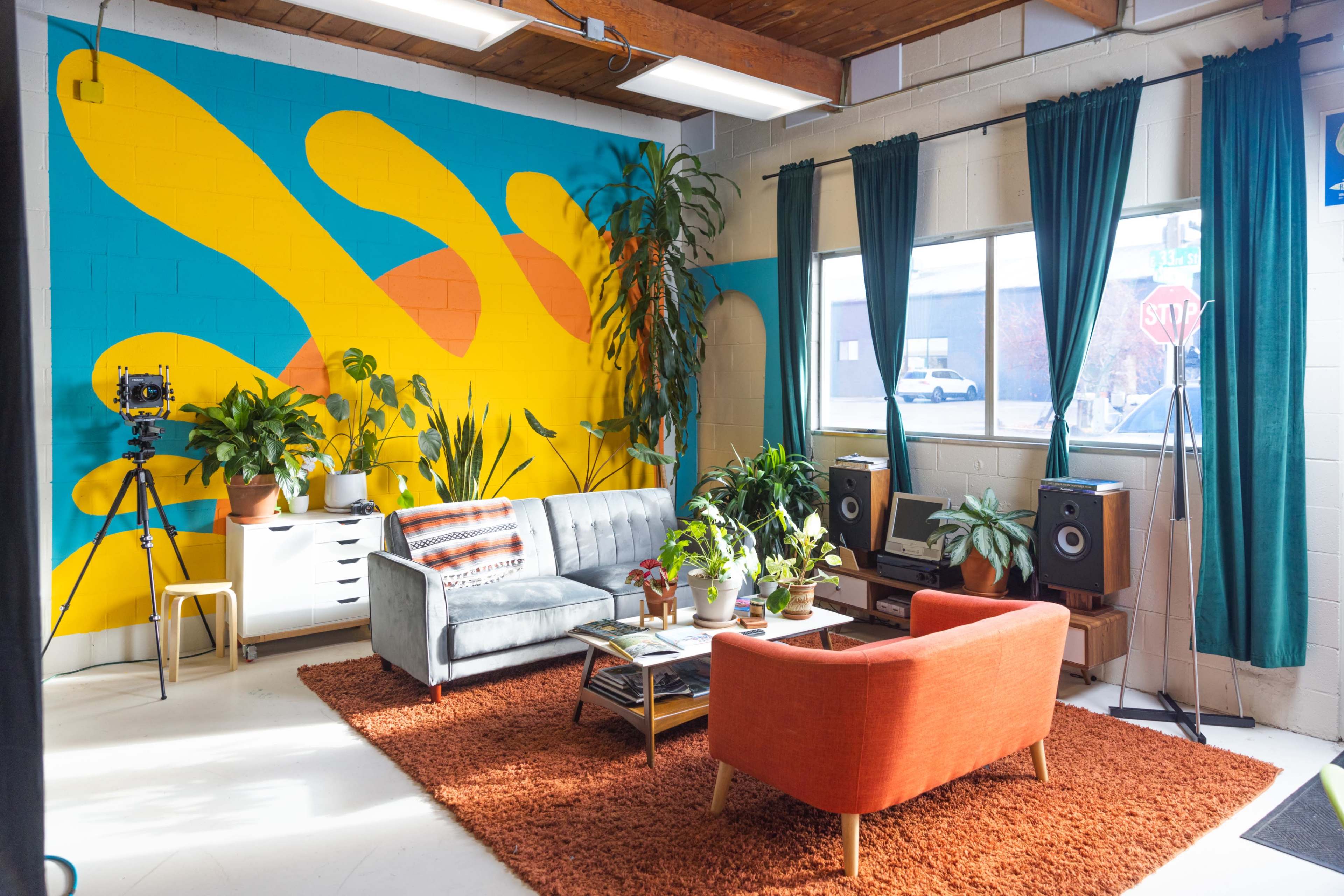 A brightly colored interior with a blue and yellow mural, featuring a gray sofa and an orange armchair, surrounded by various plants and decorative furniture.