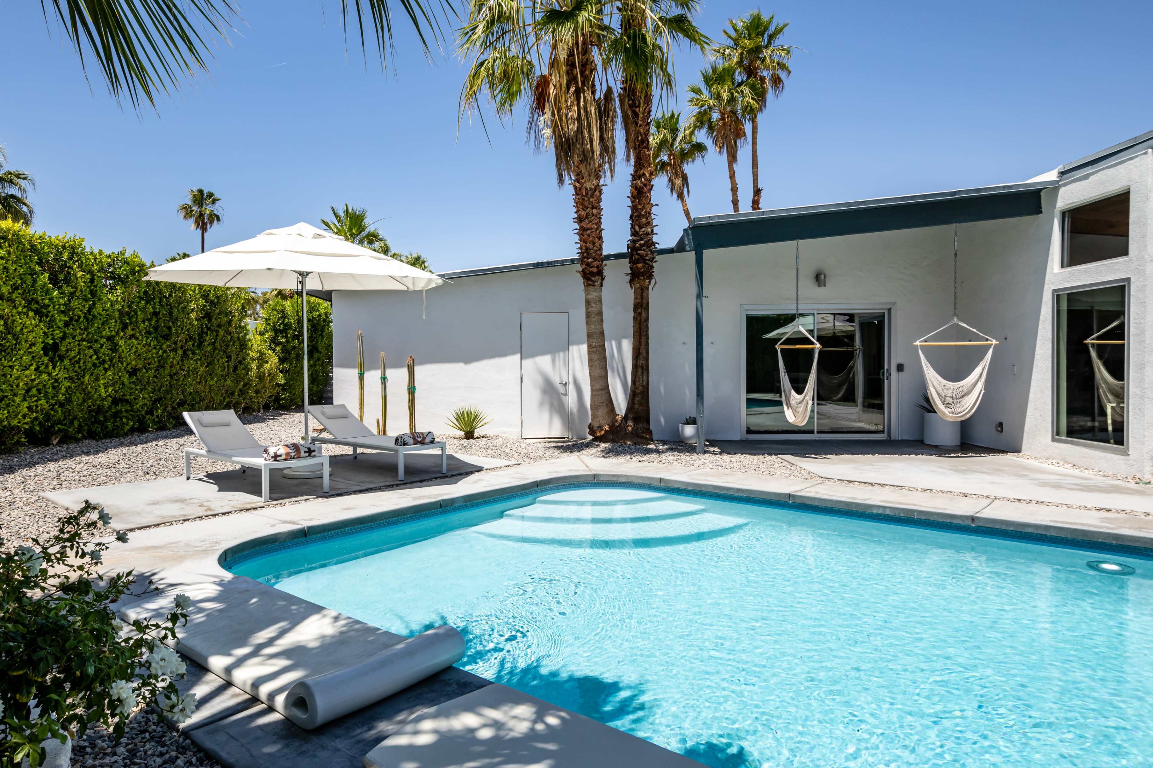 The image shows a modern swimming pool surrounded by palm trees, lounge chairs, and a hammock in a sunny outdoor setting.