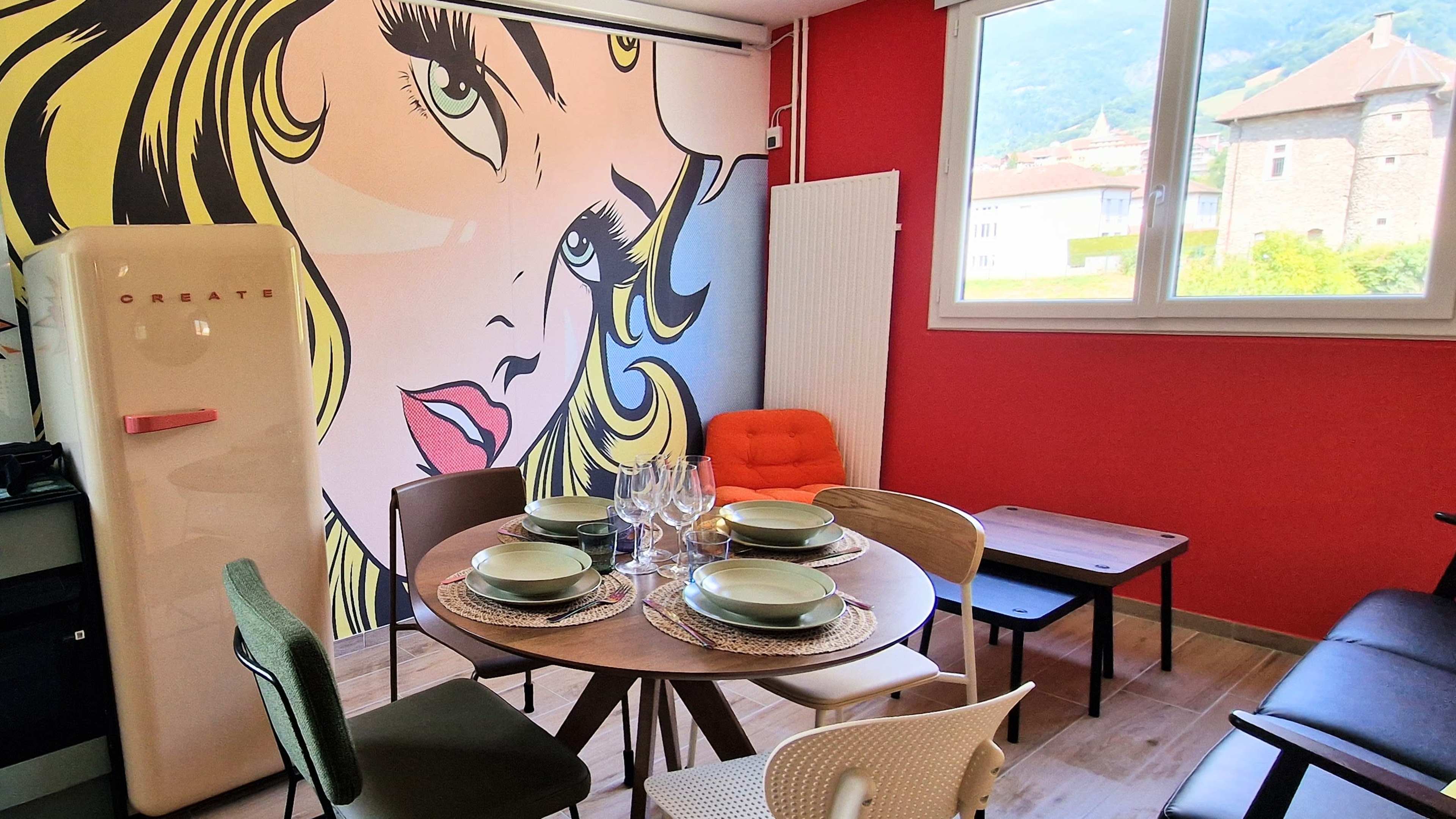 The image shows a dining area with a round table set for a meal, surrounded by various chairs, and features a large, colorful pop art mural of a woman's face on the wall.