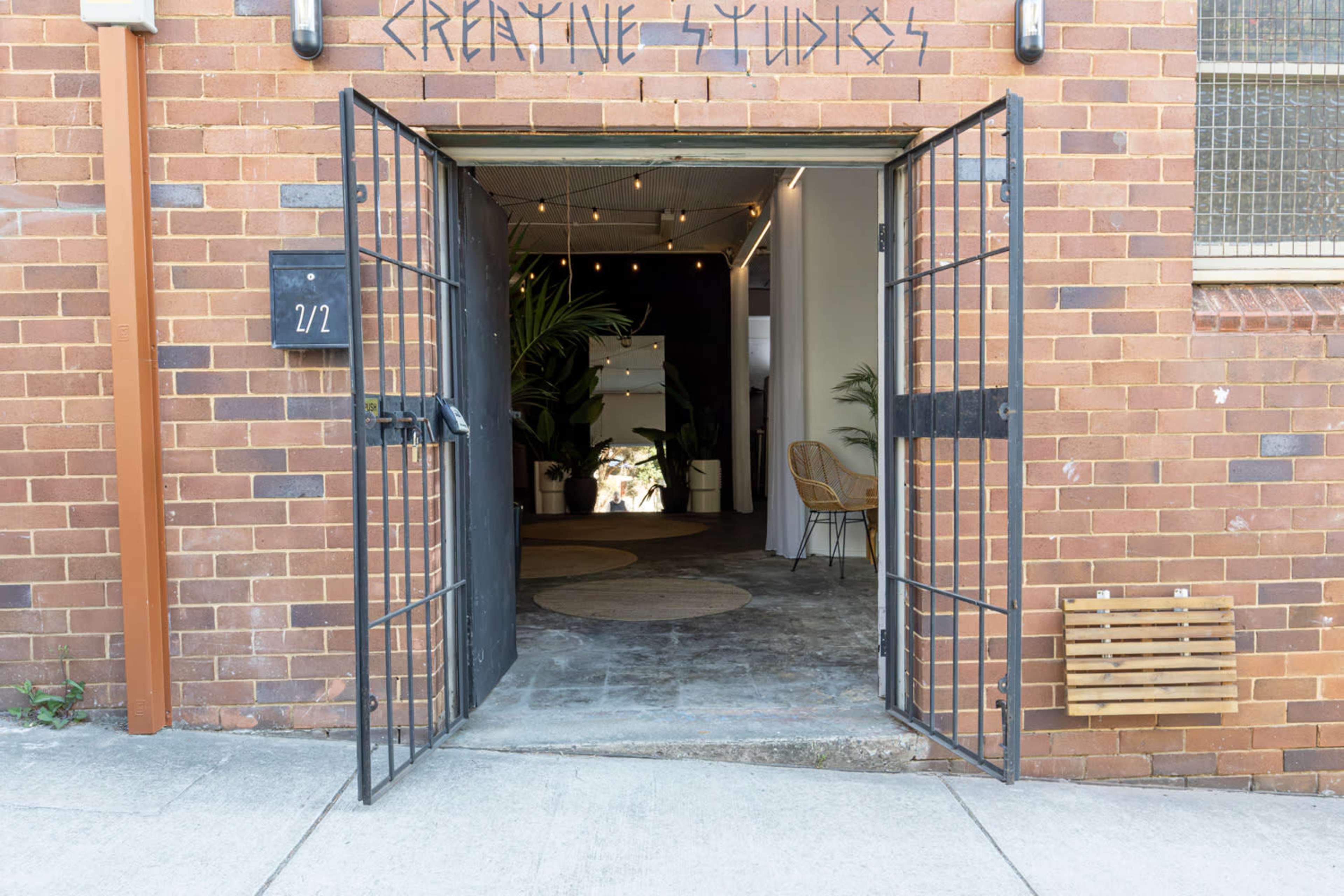 The image shows an entrance to a studio with an open iron gate and a brick wall, leading into a hallway featuring plants and seating.