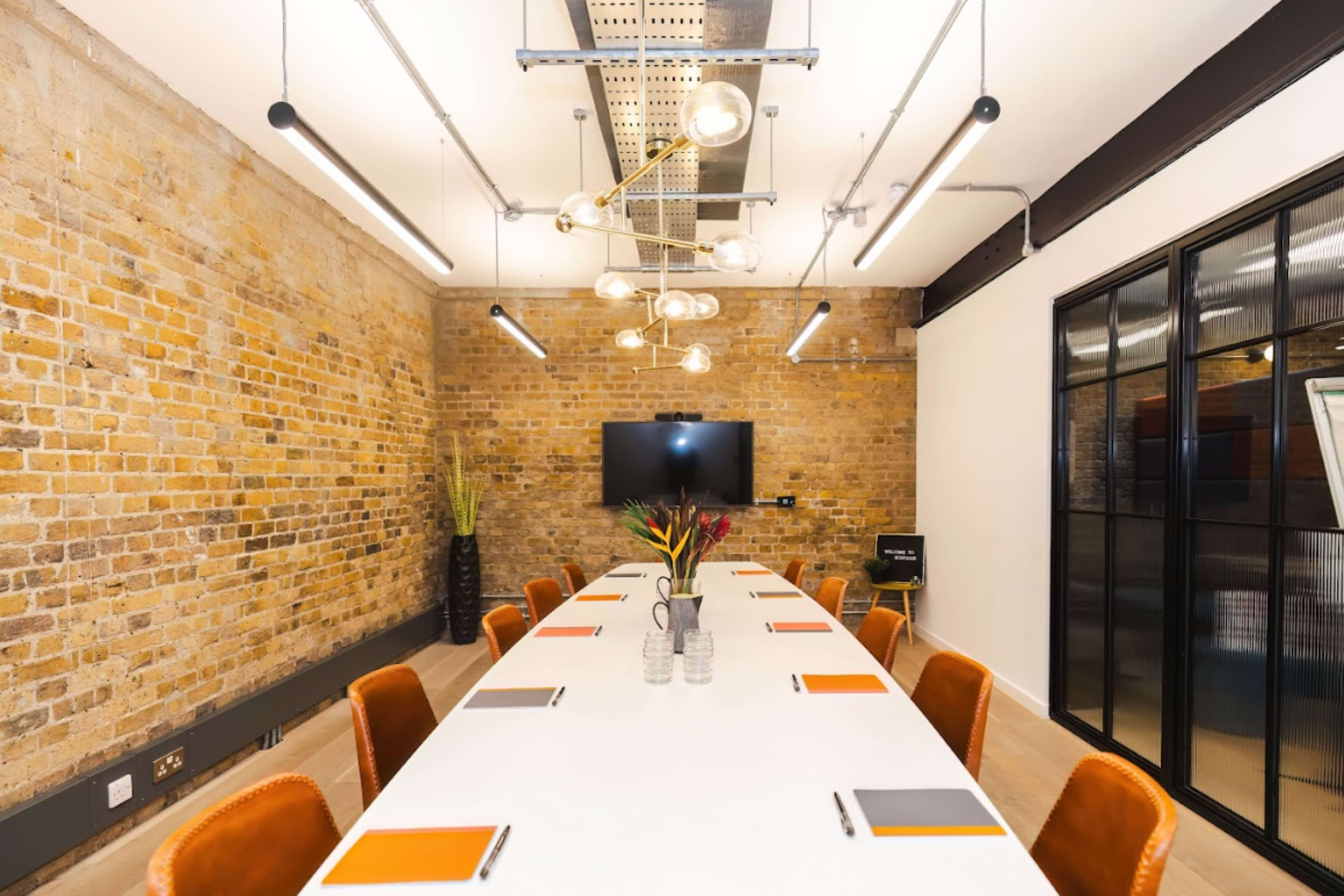 A spacious meeting room features a long white table surrounded by brown chairs, with a brick wall and modern lighting fixtures overhead.