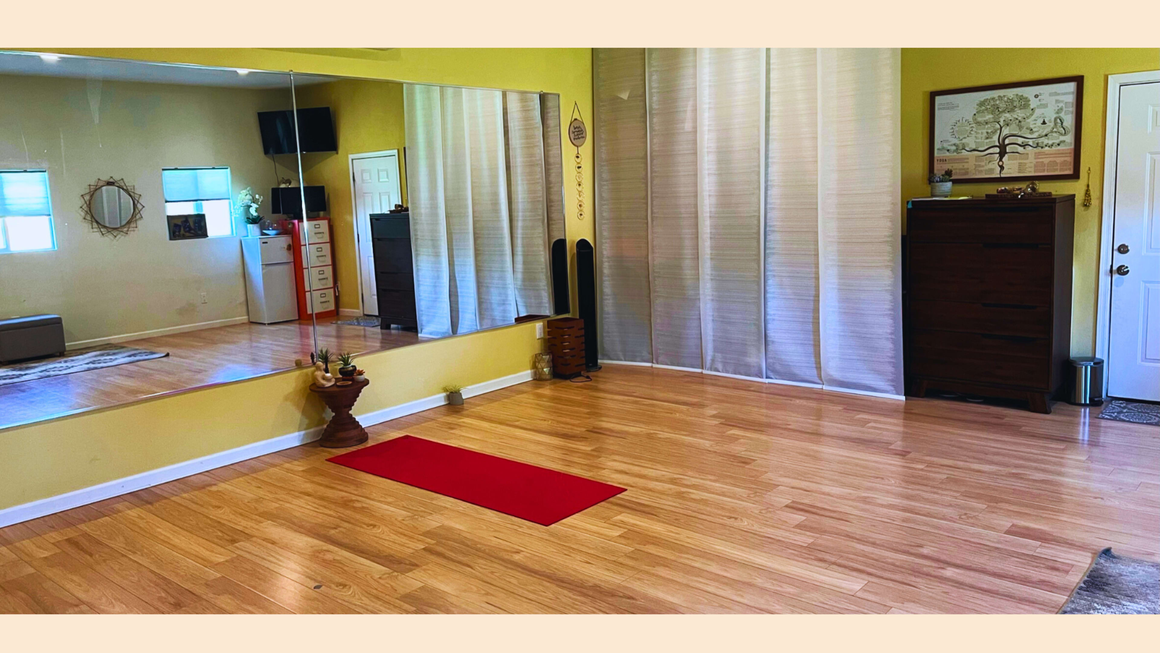 A spacious room with wooden flooring, a large mirror on one wall, a red yoga mat on the floor, and a wooden dresser near a door.