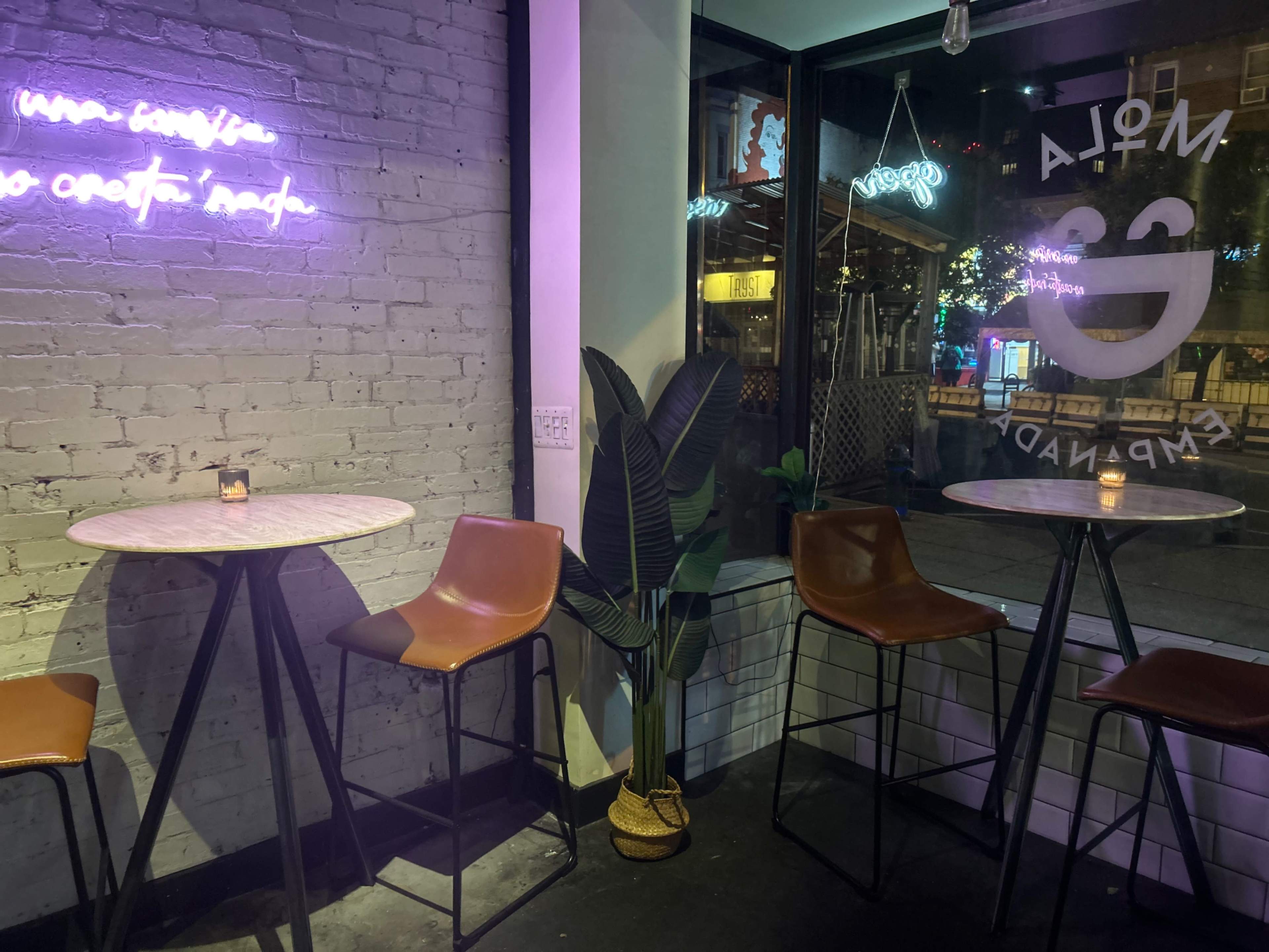 The image shows a modern interior of a café with tall seating at small tables, a large window displaying street views, and neon lighting on a brick wall.