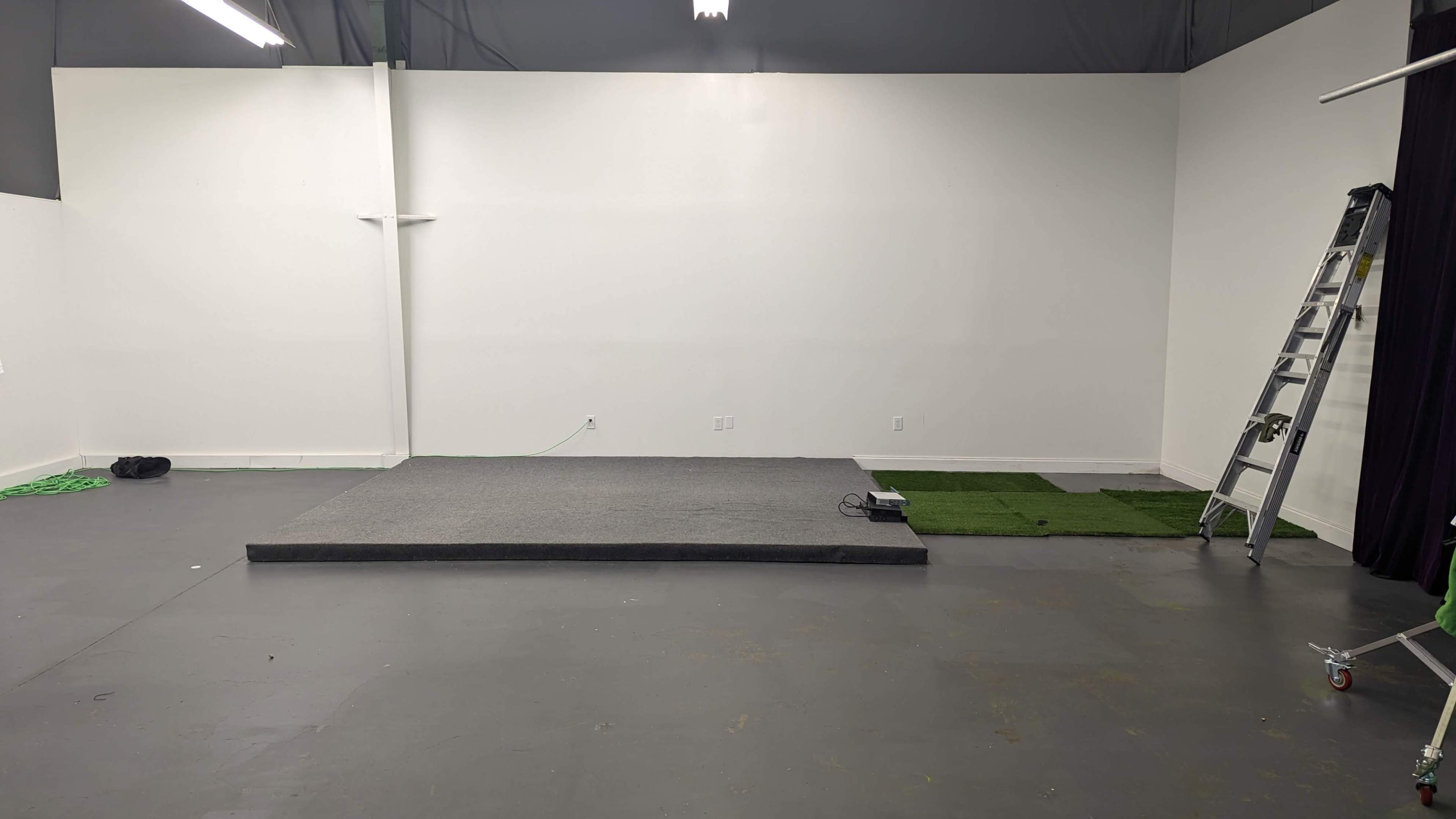A gray stage platform is positioned against a blank white wall in a spacious, empty room with a small patch of green turf nearby.