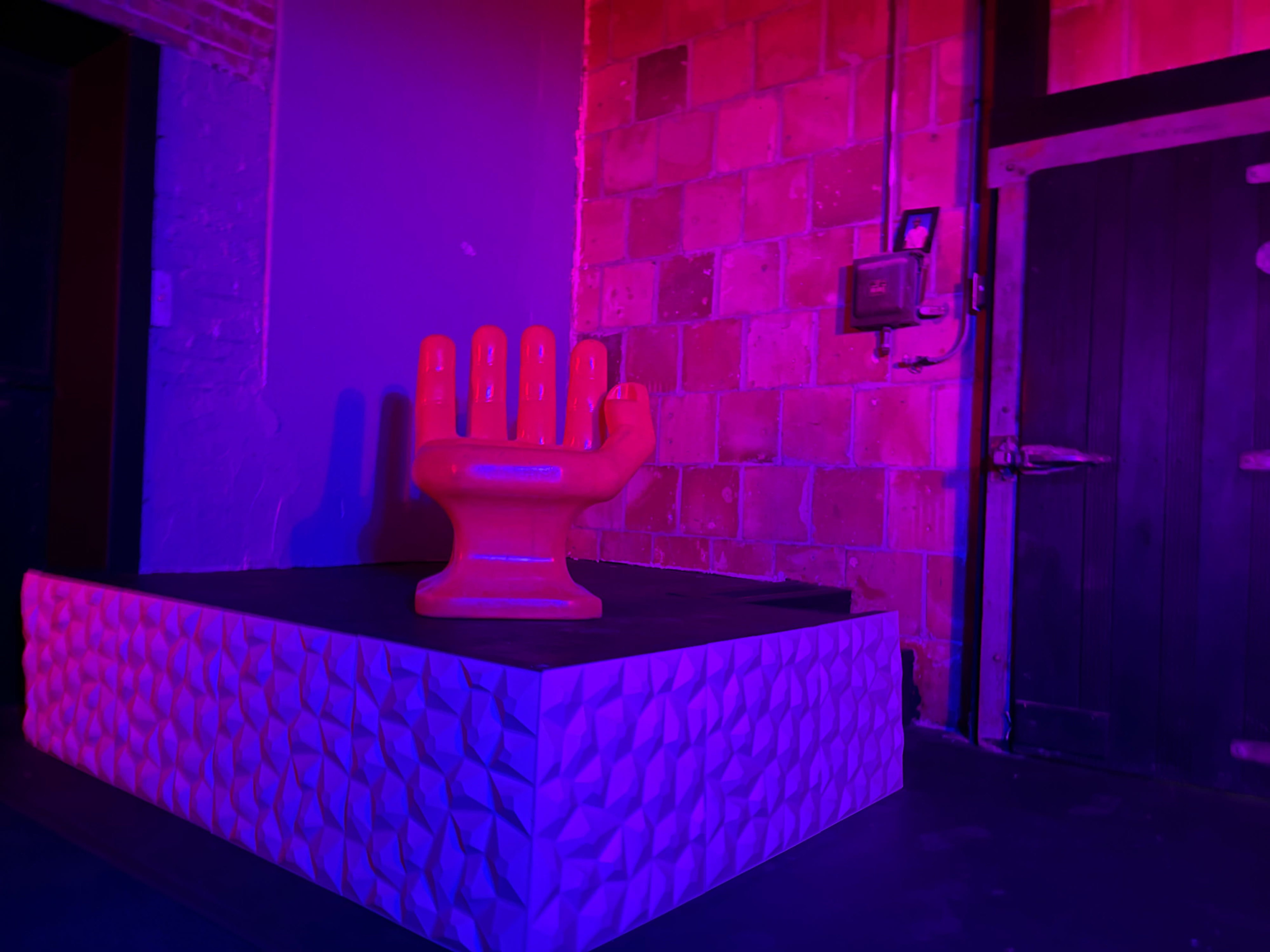 A vibrant red hand sculpture is displayed on a textured white pedestal against a backdrop of colorful lighting and a brick wall.
