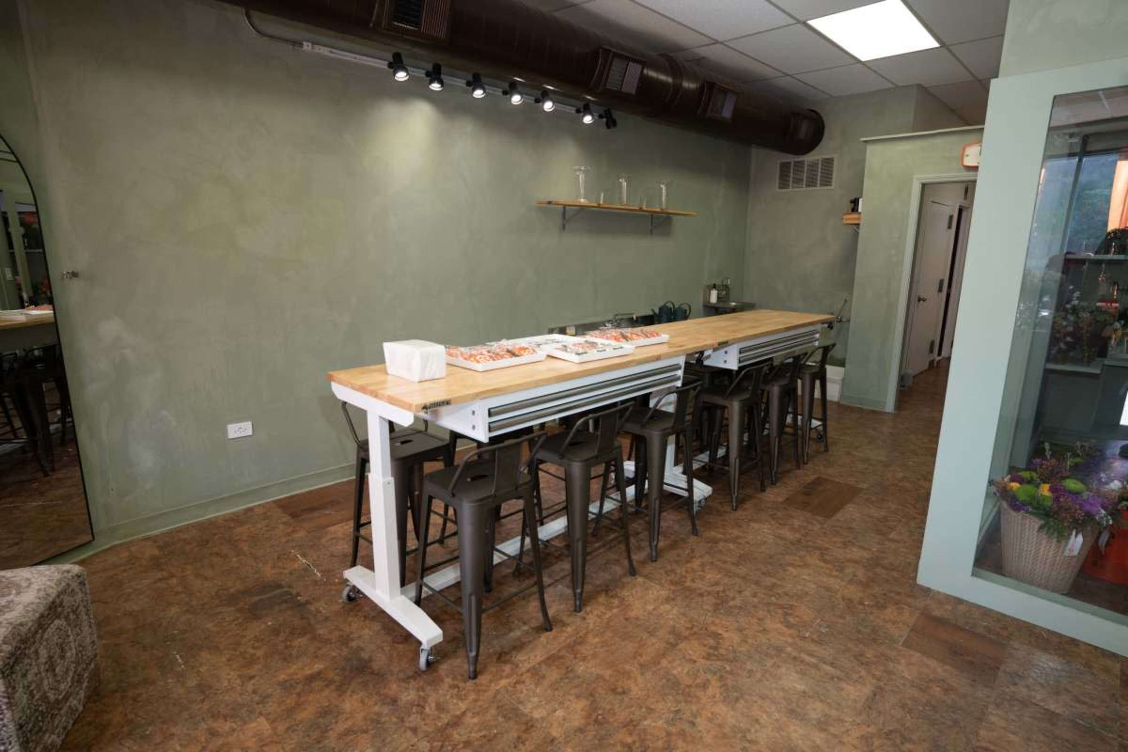 The image shows a spacious, unfinished room featuring a long wooden table with metal stools, alongside a greenish wall and various fixtures.
