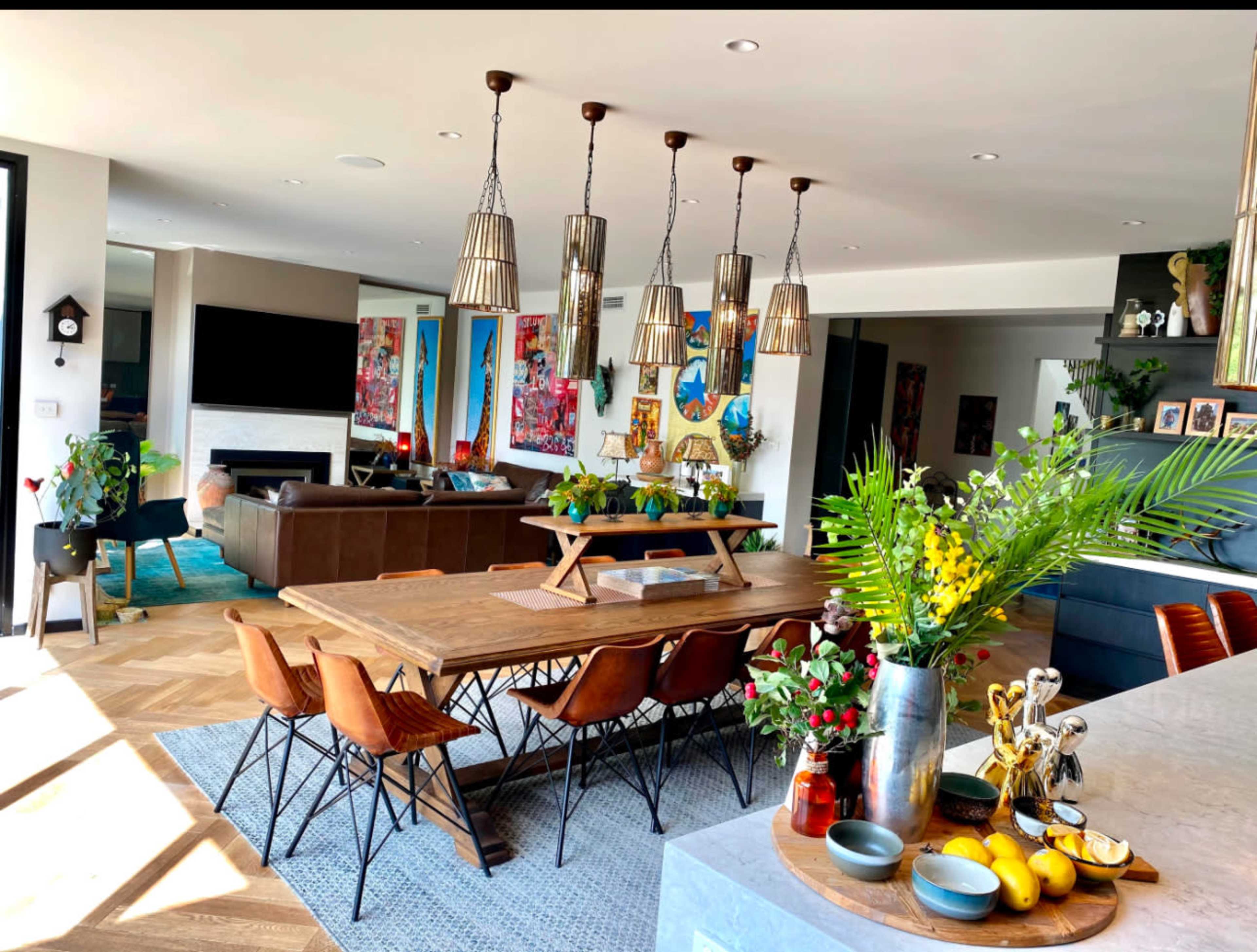 A spacious dining area features a large wooden table surrounded by stylish chairs, with pendant lights hanging above and a kitchen partially visible in the background.
