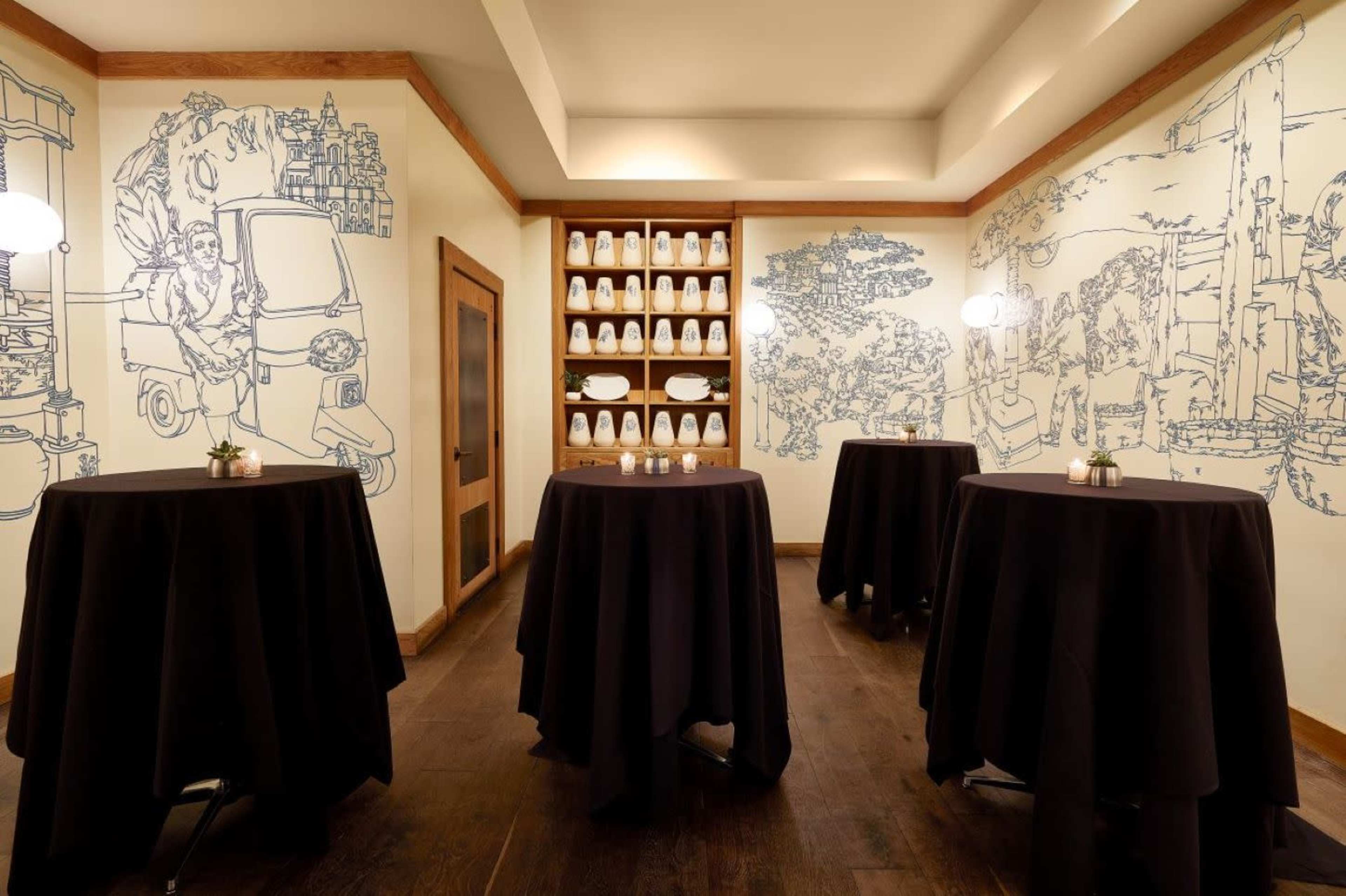 The image shows a small dining area with tall tables covered in black tablecloths, surrounded by walls featuring blue illustrations and a shelf filled with white dishware.