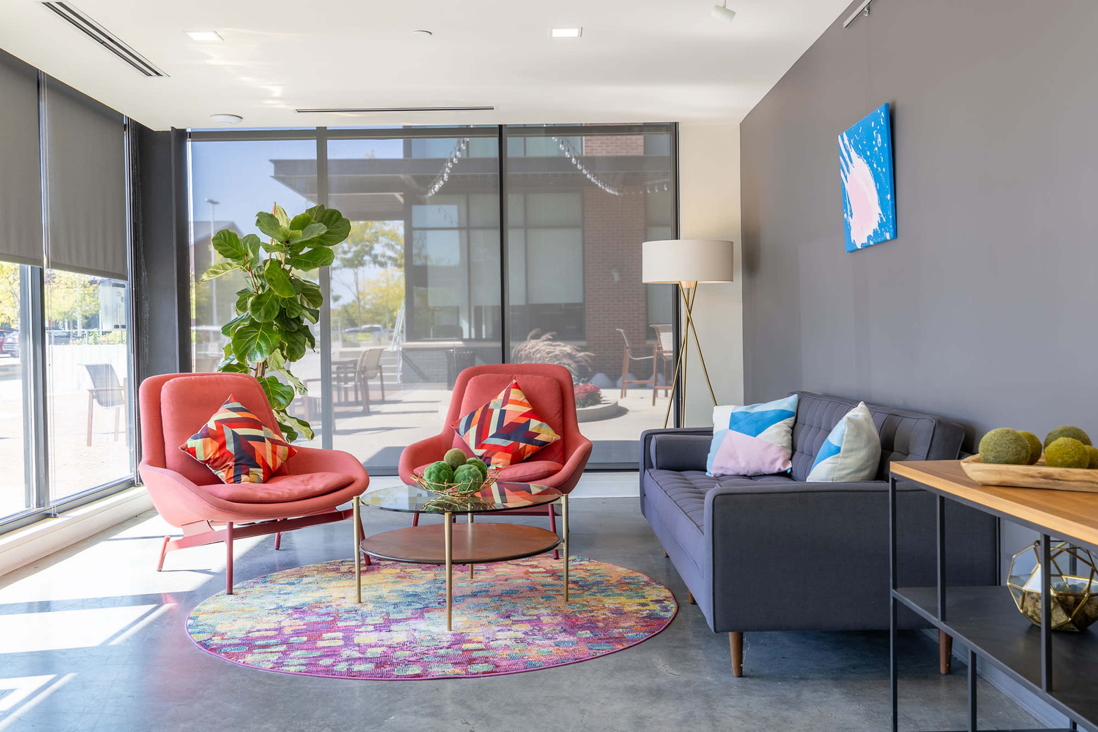 The image shows a modern lounge area featuring two pink chairs, a gray sofa, a round glass coffee table, and a vibrant area rug, all set against large windows that provide natural light.