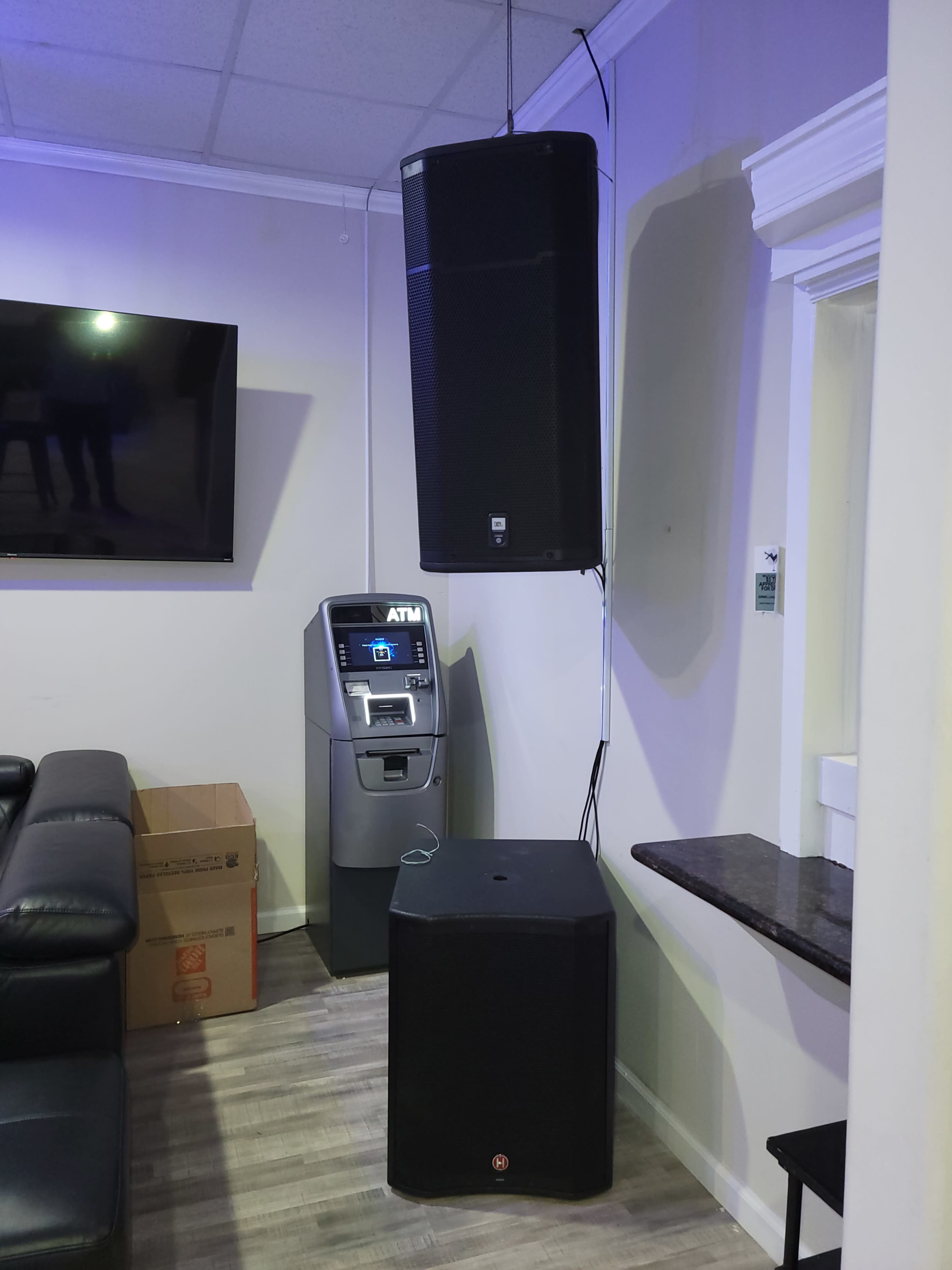 Two large speakers mounted on a wall next to an ATM in a room with a leather couch and a countertop.