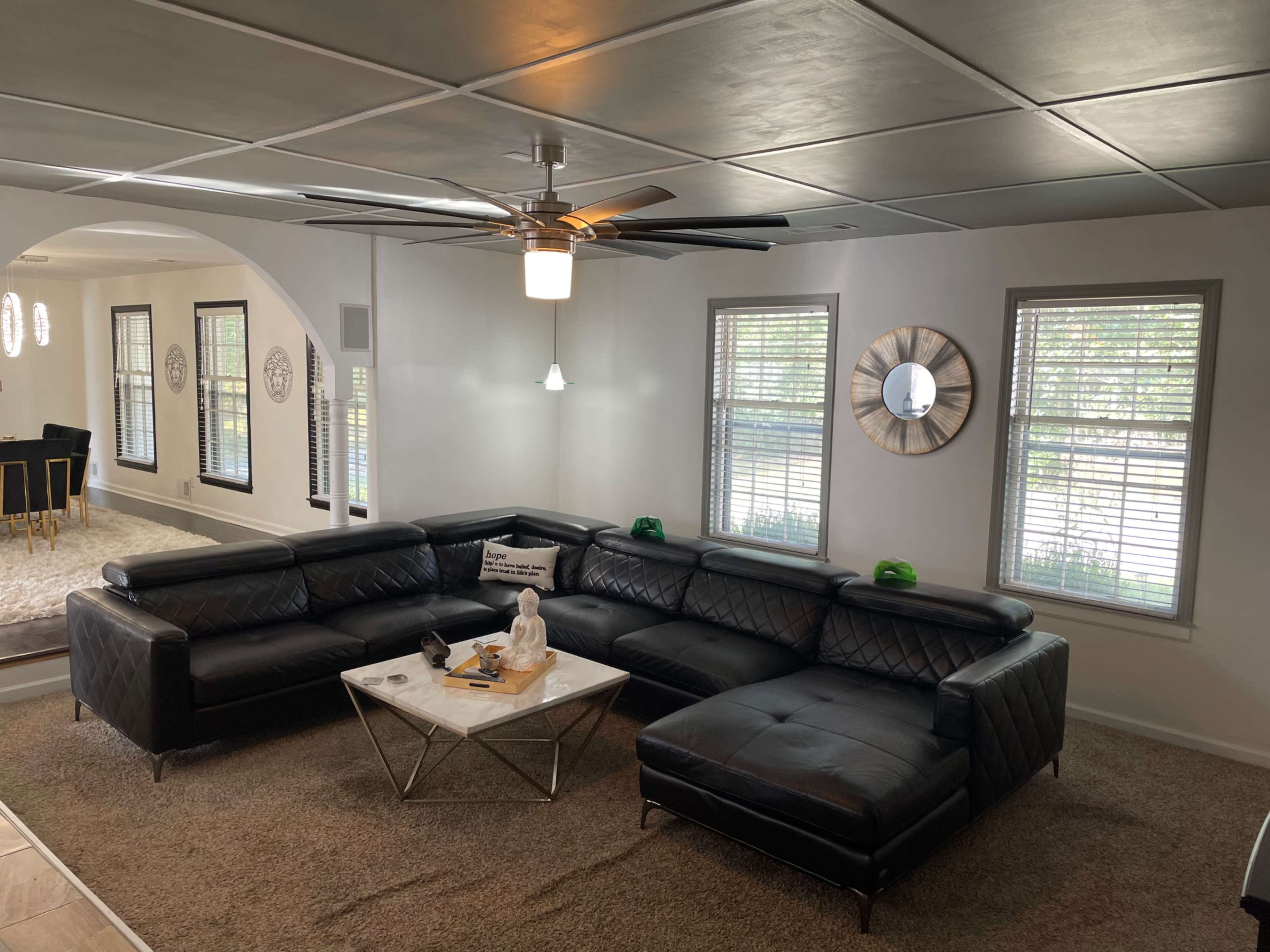 A modern living room features a large black sectional sofa, a white coffee table, and several windows adorned with blinds.