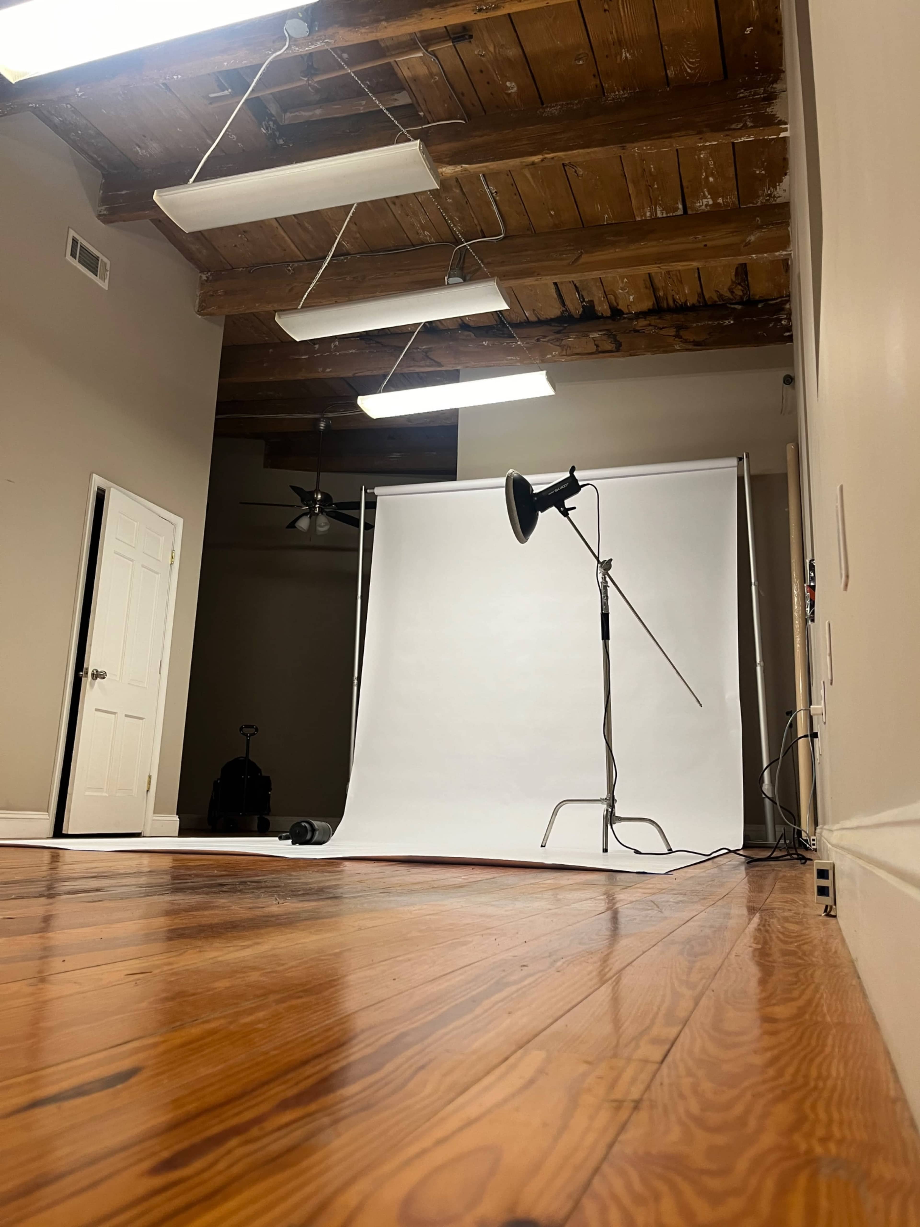 The image shows a photography studio with a white backdrop, a light stand, and wooden floors.
