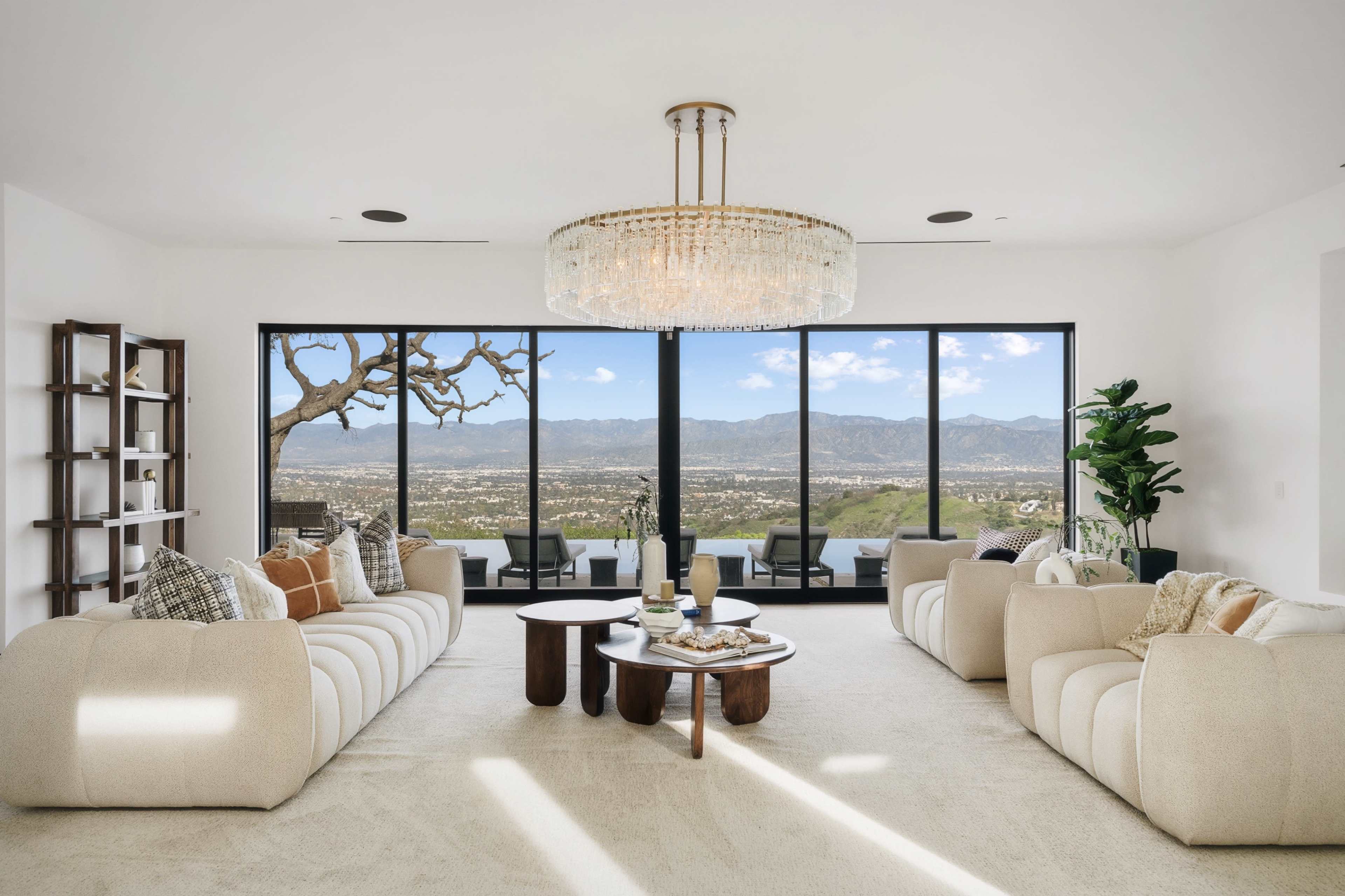 A modern living room features two cream-colored sofas, a wooden coffee table, and large windows that showcase a panoramic view of the mountains and valley beyond.