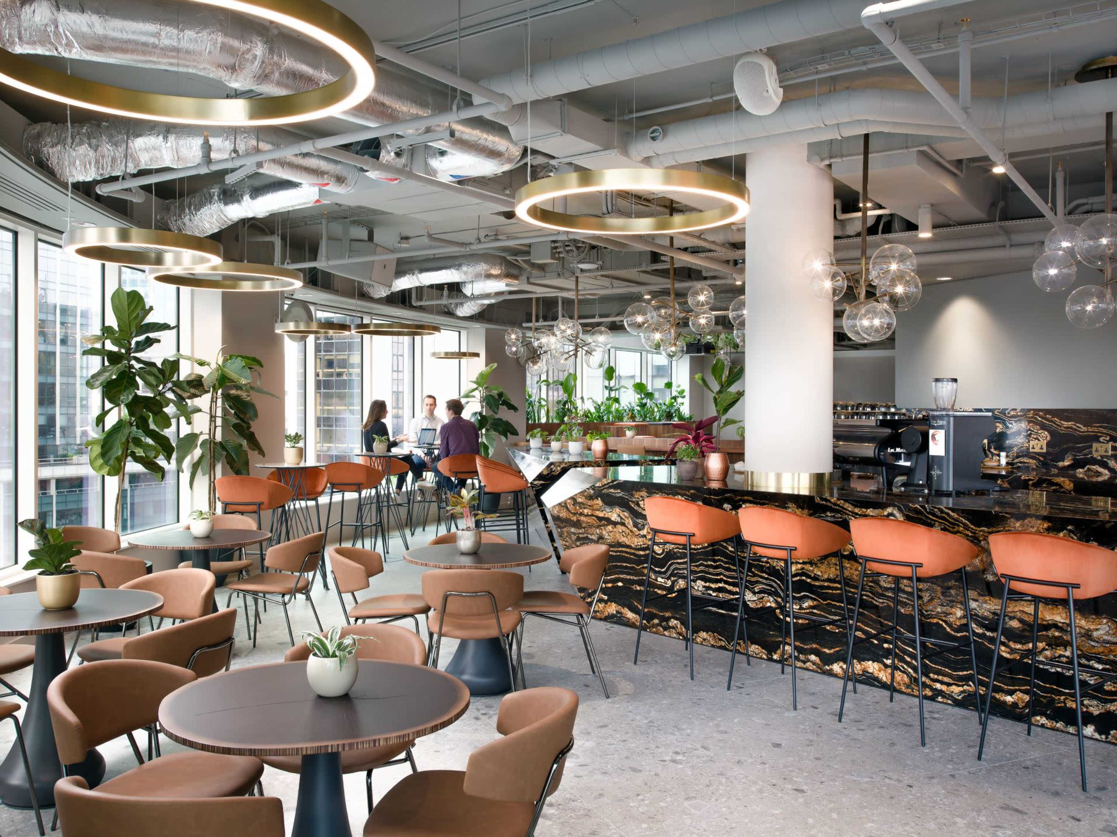 The image shows a modern cafe interior with circular tables, stylish seating, and a marble counter, accented by hanging light fixtures and greenery.
