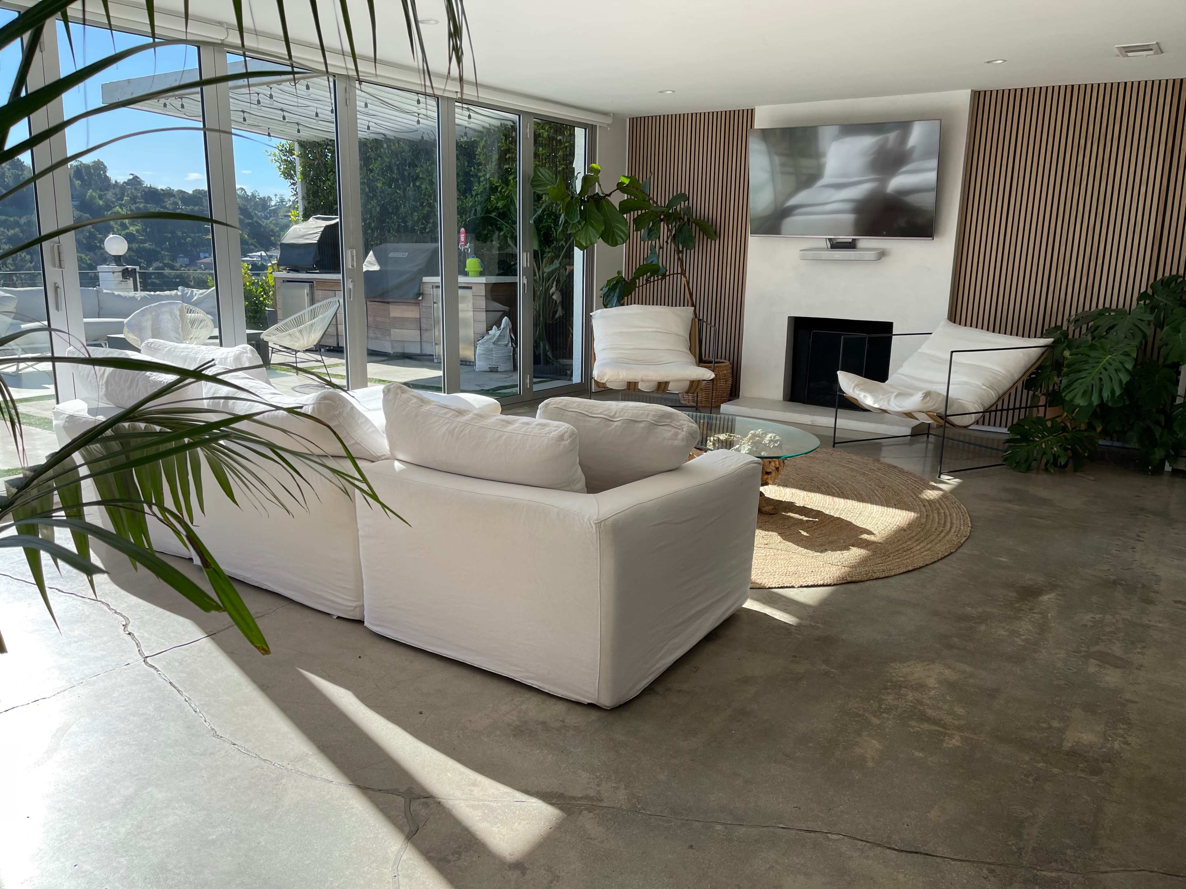 The living room features a white sofa set, a round coffee table, and large windows that provide natural light, with plants and a television visible in the background.