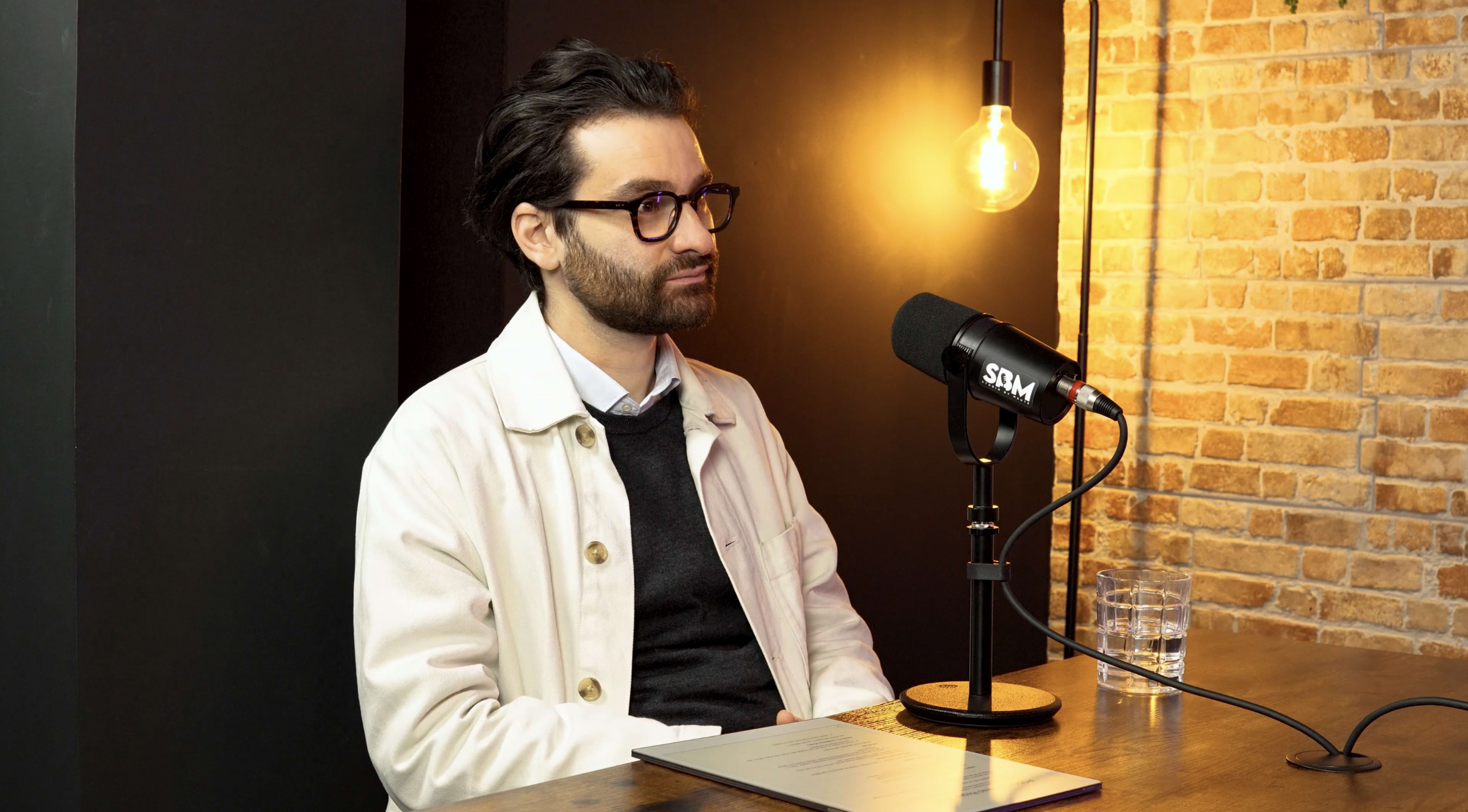A man with glasses sits at a table in a dimly lit room, positioned in front of a microphone and a glass.