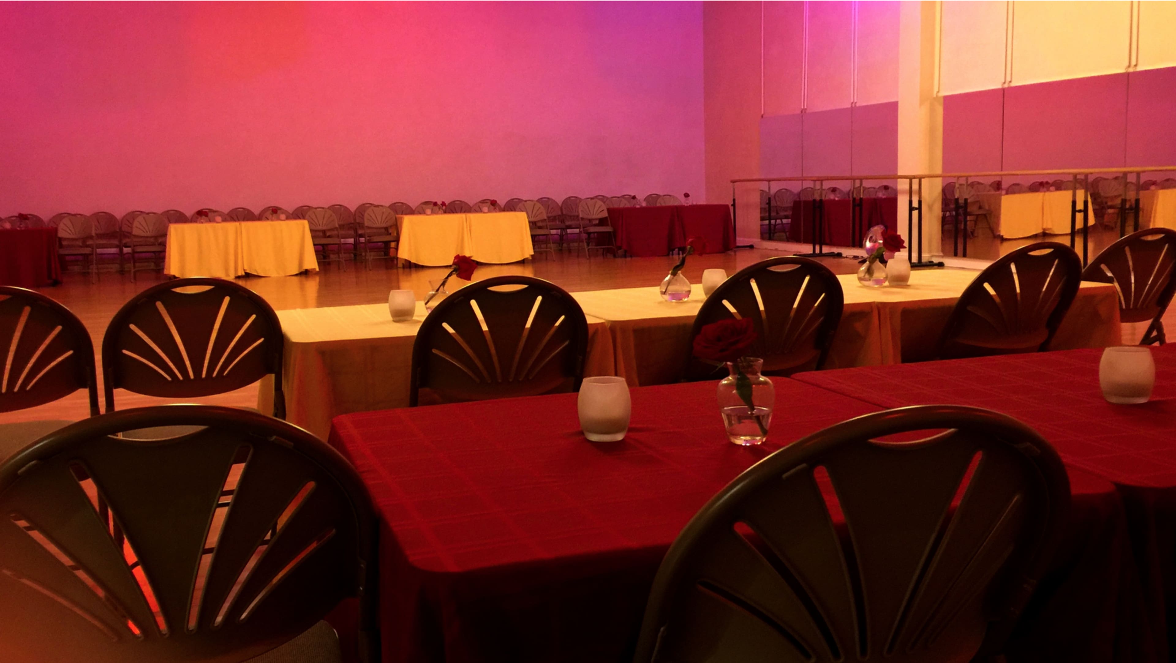 A dance studio with tables set in rows, some covered with red and yellow tablecloths, and chairs arranged around them.