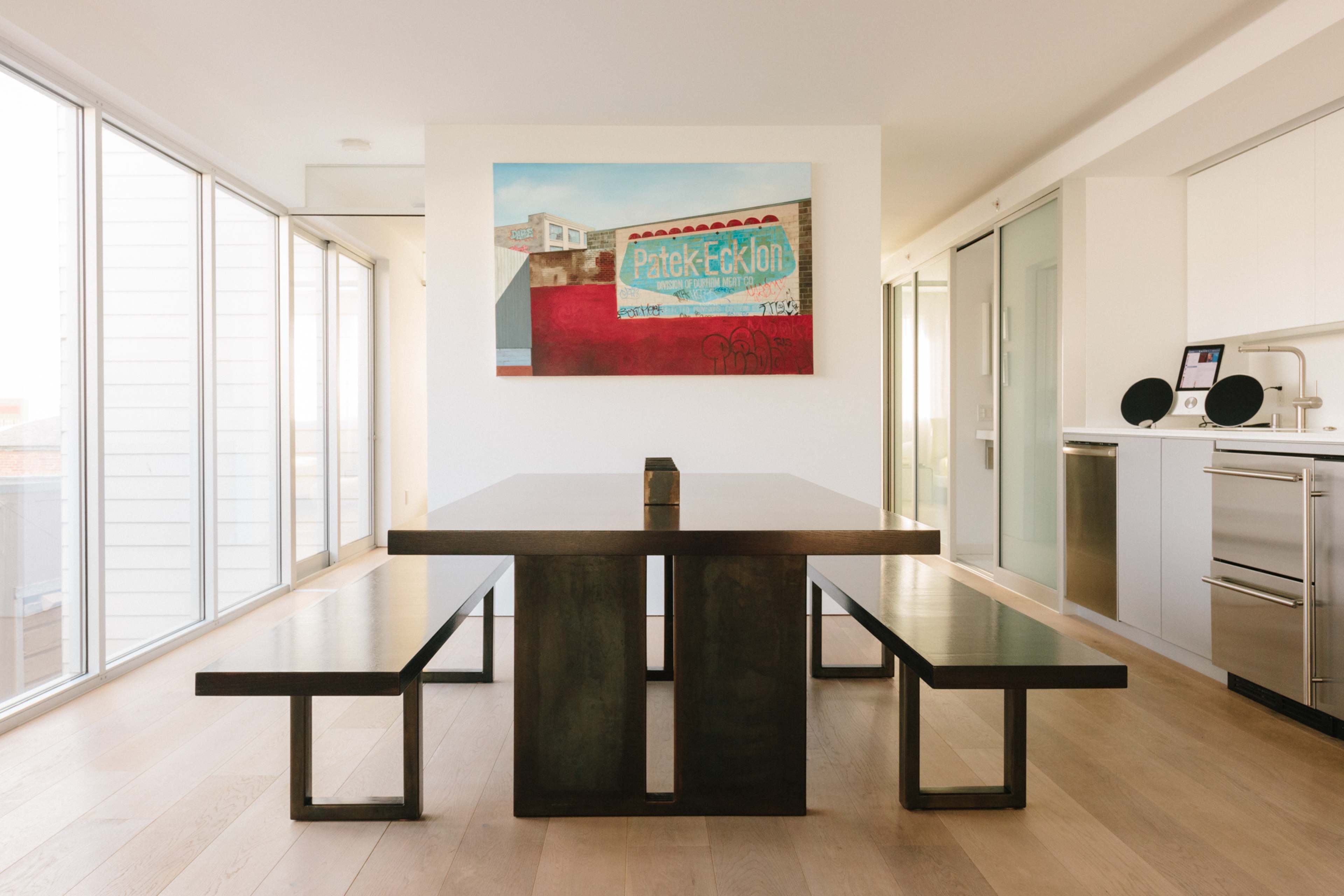 A minimalist dining area features a long dark table with matching benches, framed by large windows and a colorful artwork on the wall.