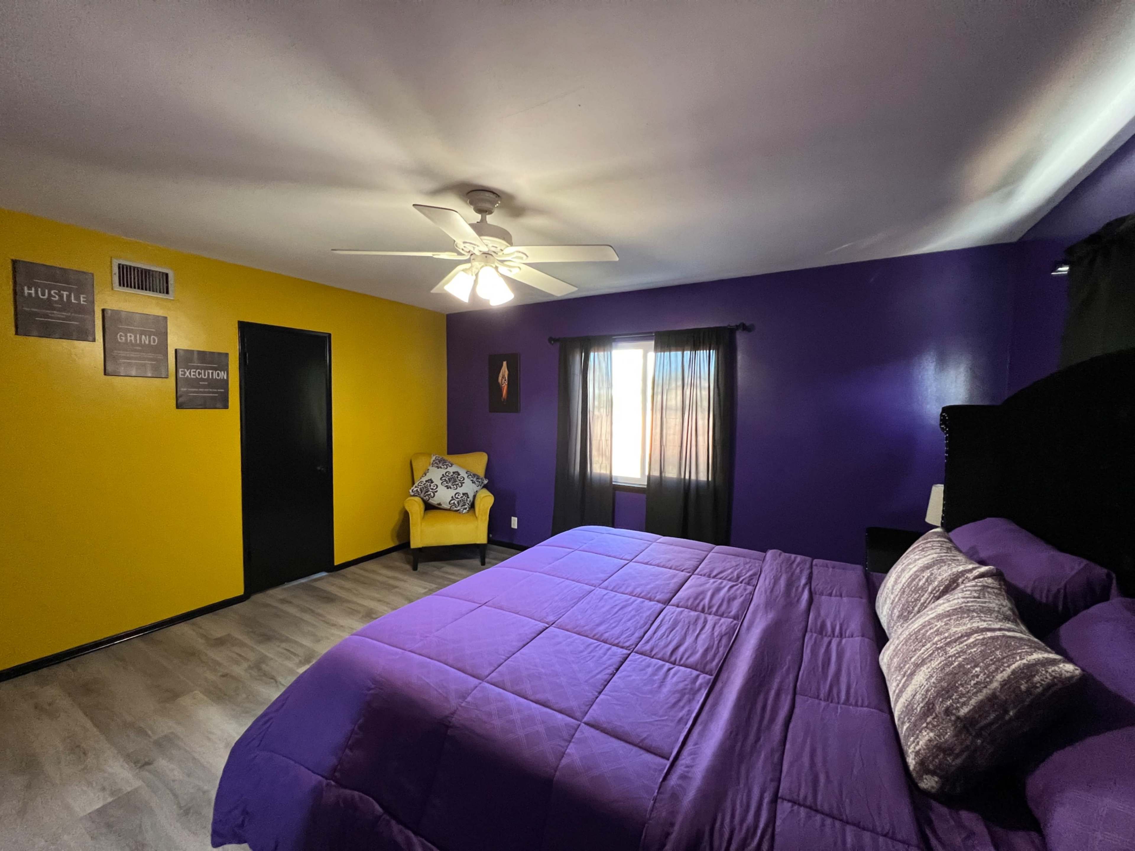 The room features a purple and yellow color scheme with a large bed, a ceiling fan, and a chair in the corner.