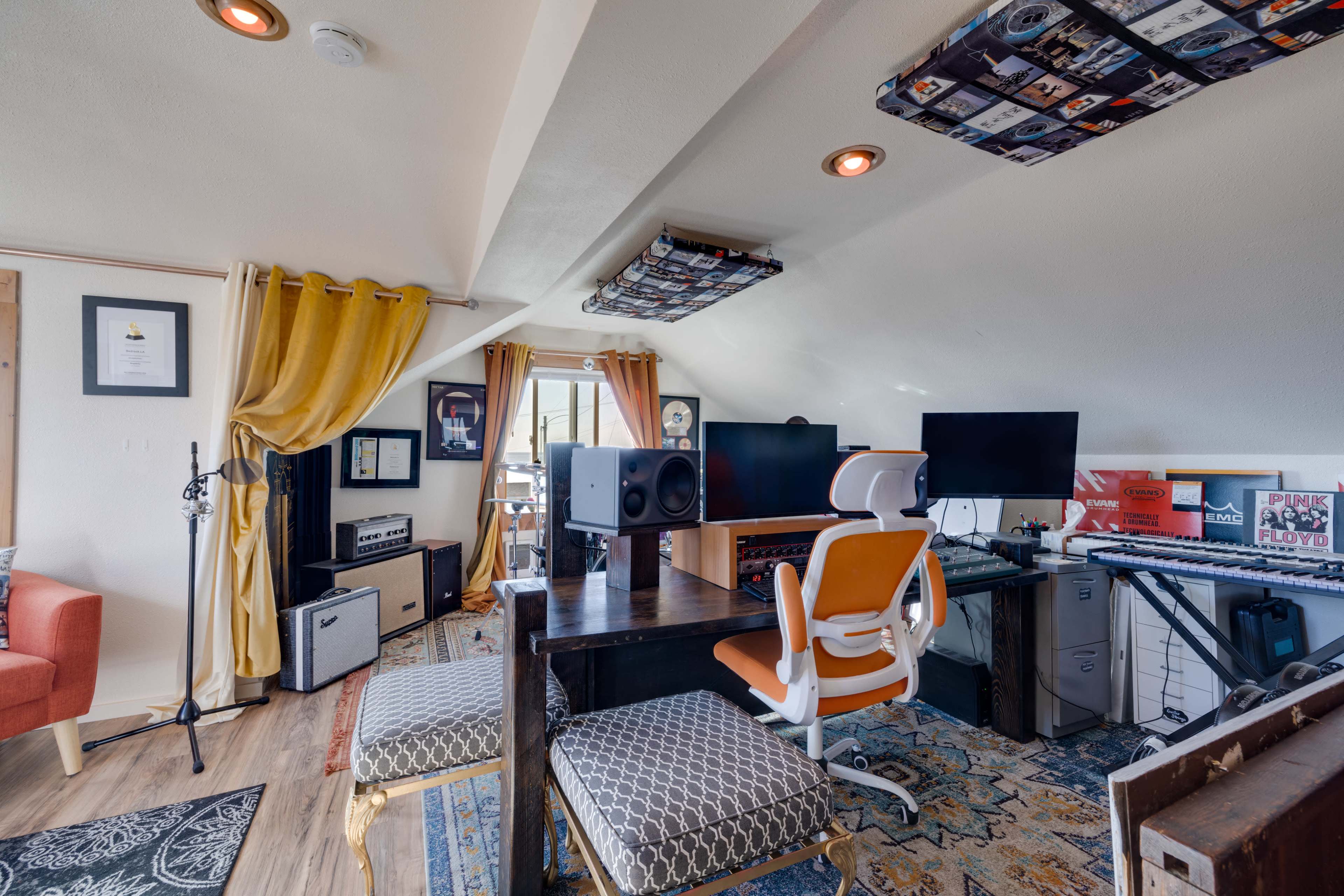 The image shows a stylish home studio with a desk, monitors, a keyboard, and various musical equipment in a bright, organized space.