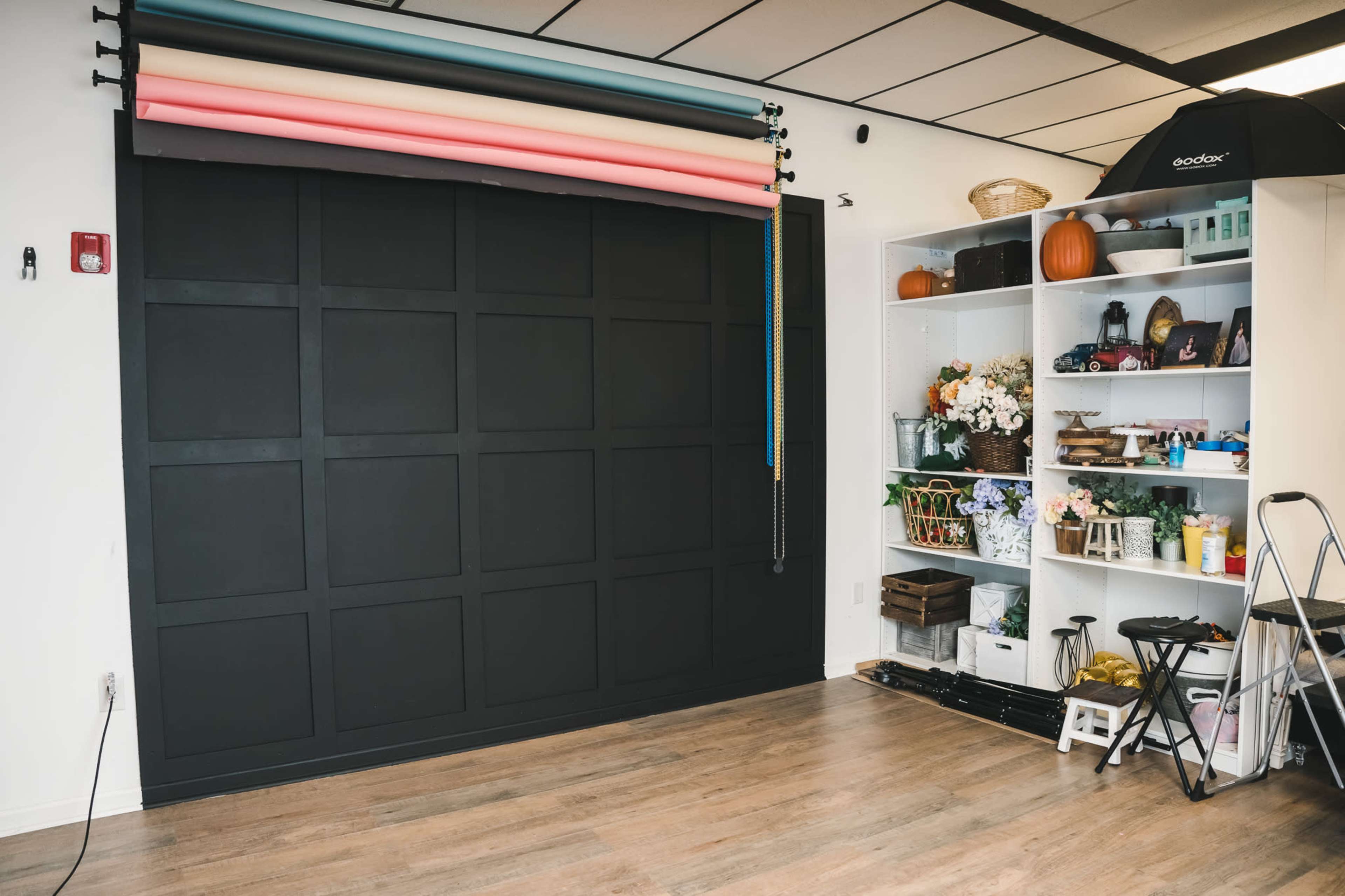 The image shows a photography studio with a black backdrop, a multi-colored roll of paper, and a shelf filled with props and decorations.