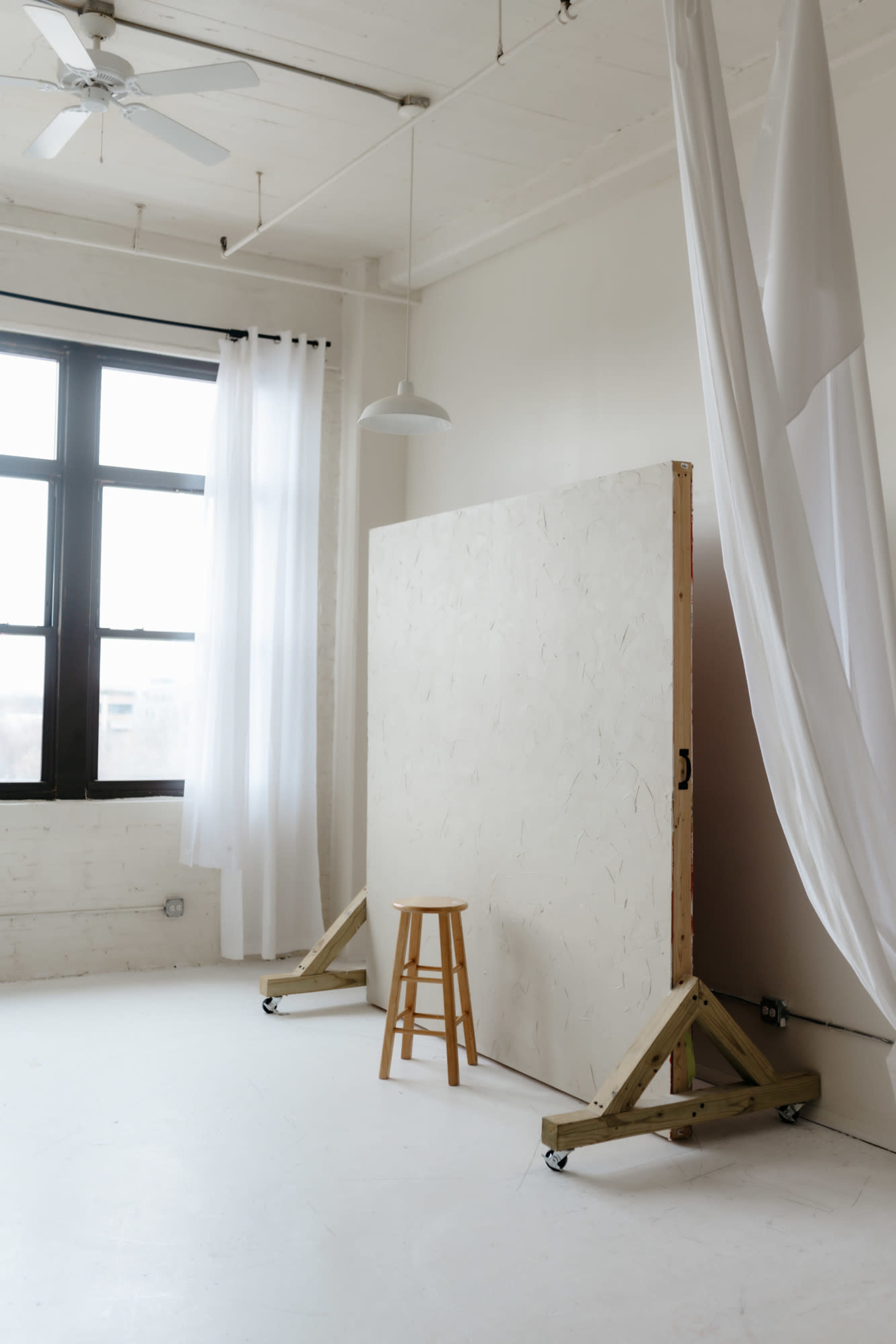 The image shows a minimalist studio space with a large white panel, a wooden stool, and large windows letting in natural light.