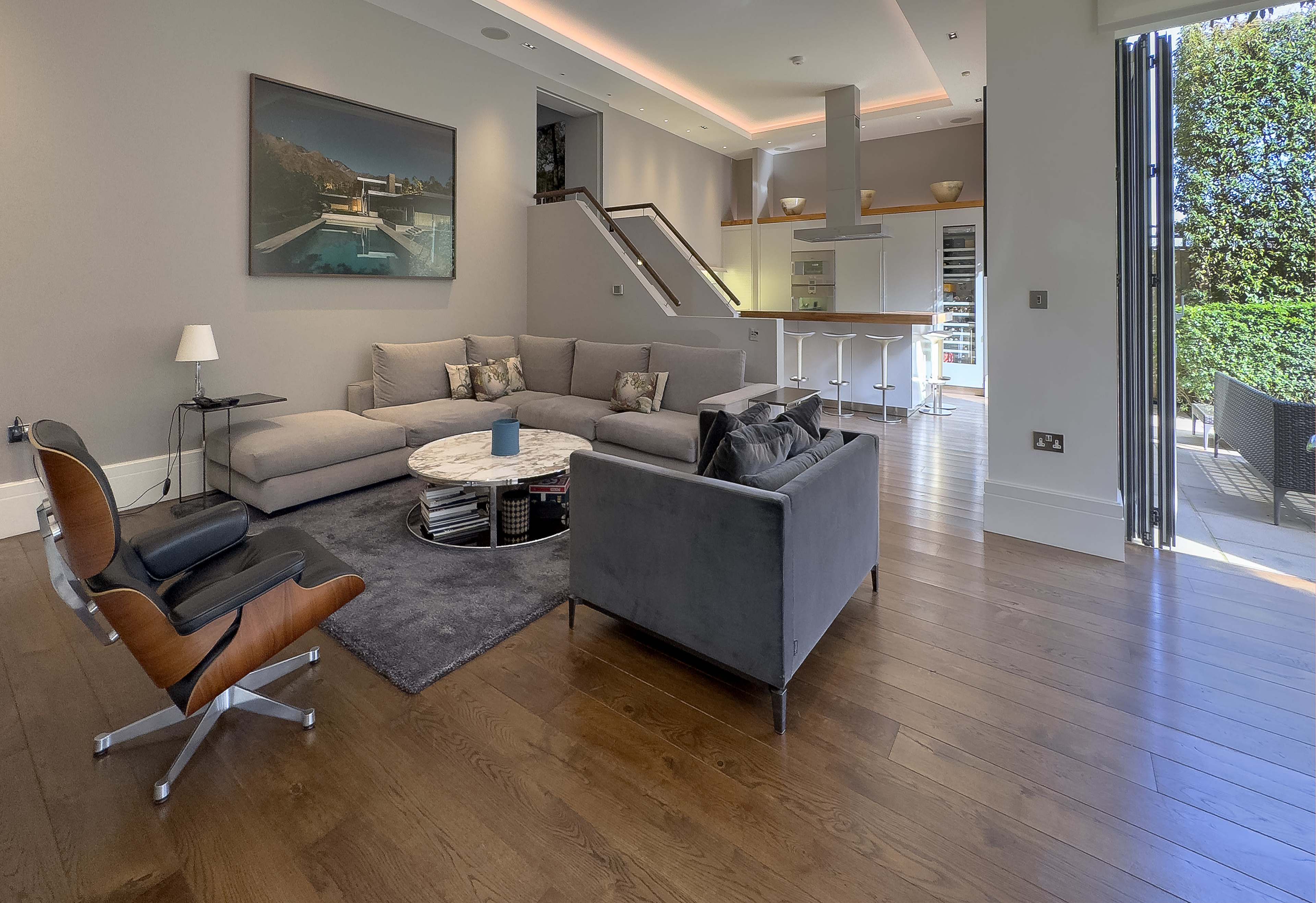 The image shows a modern living room featuring a large sectional sofa, a round coffee table, and a staircase leading to an upper level, with large windows providing natural light.