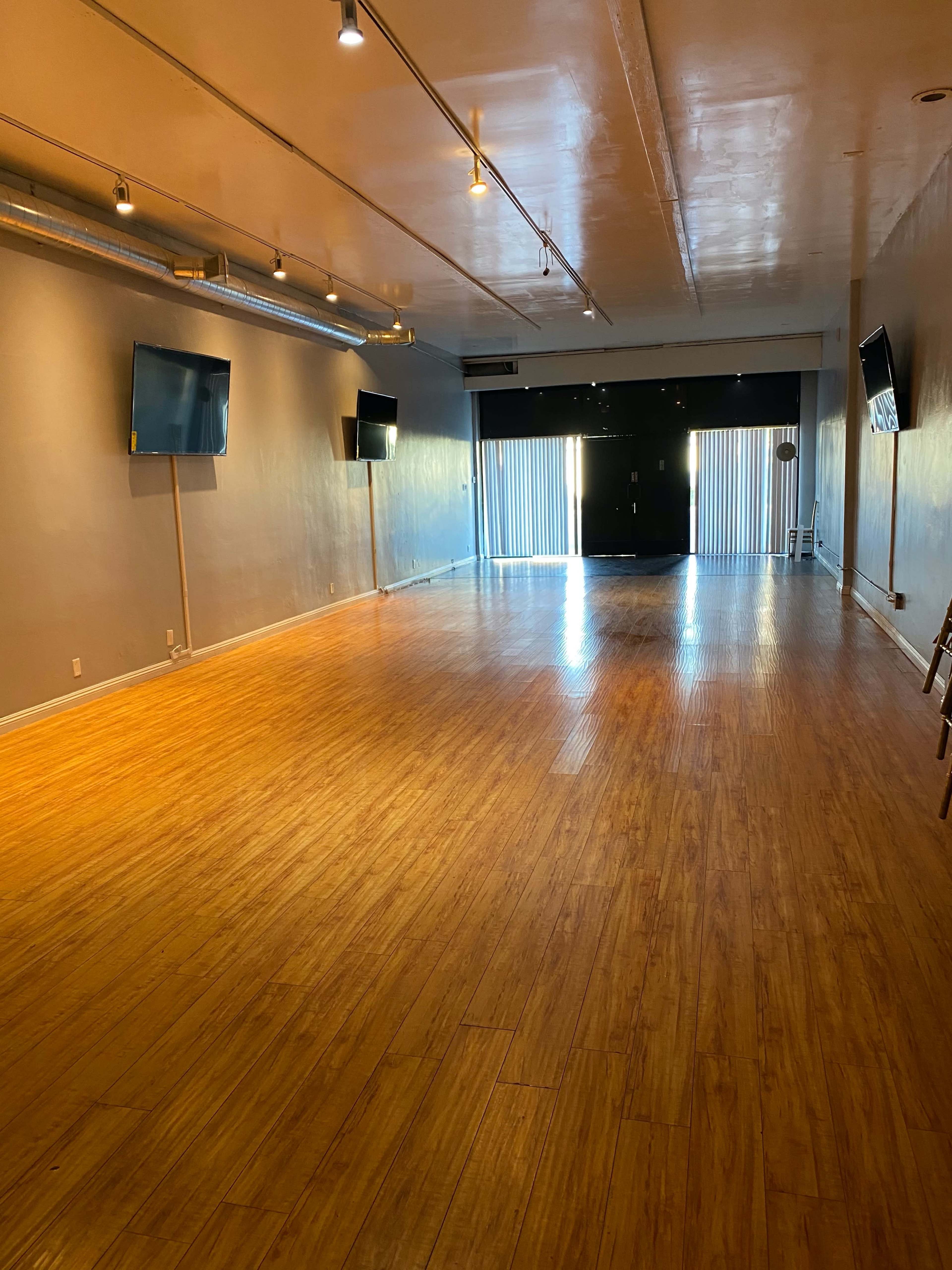The image shows an empty studio space with wooden flooring, two mounted televisions on the walls, and large windows covered by blinds at one end.