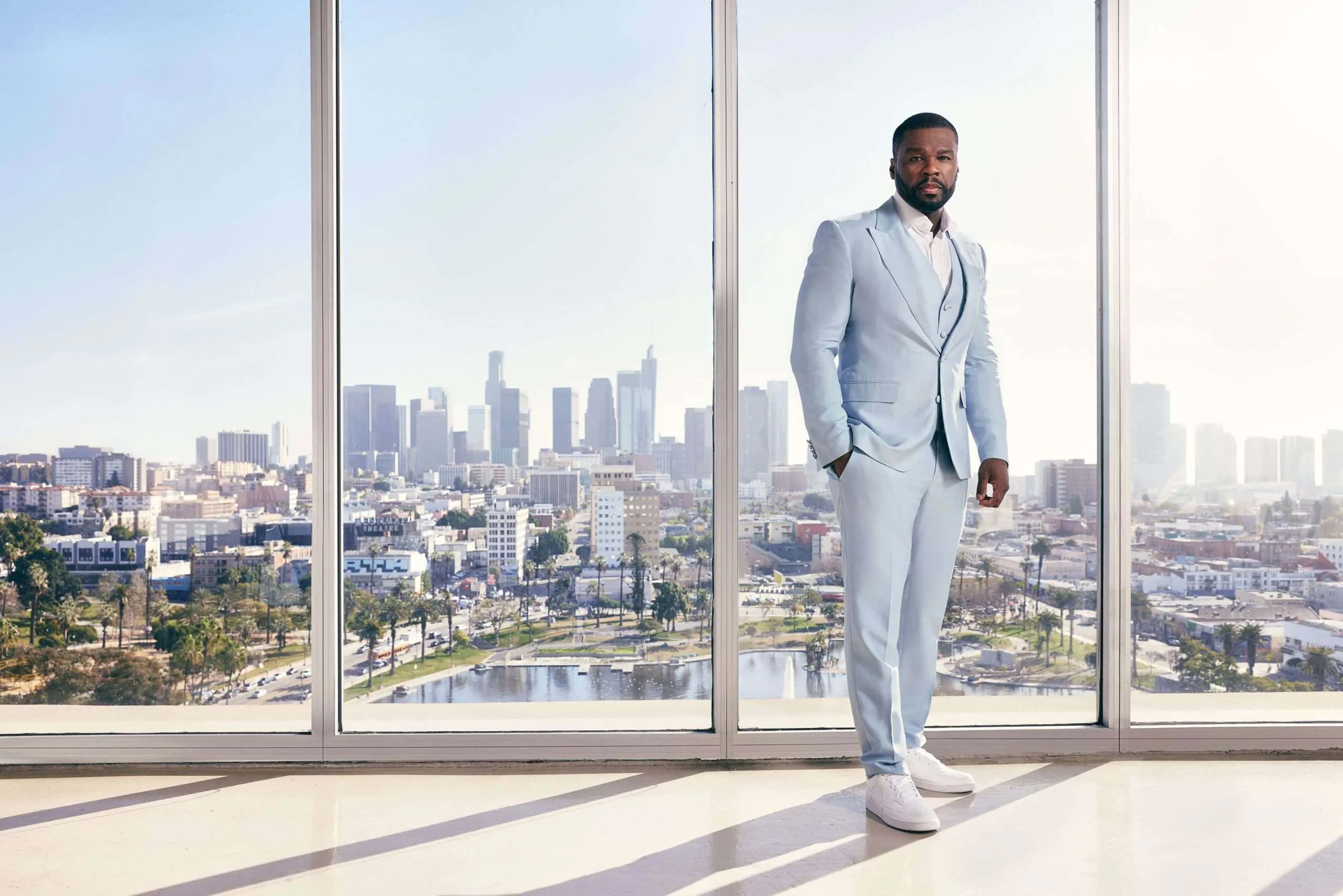 A man wearing a light blue suit and white sneakers stands in front of a large window with a view of a city skyline.