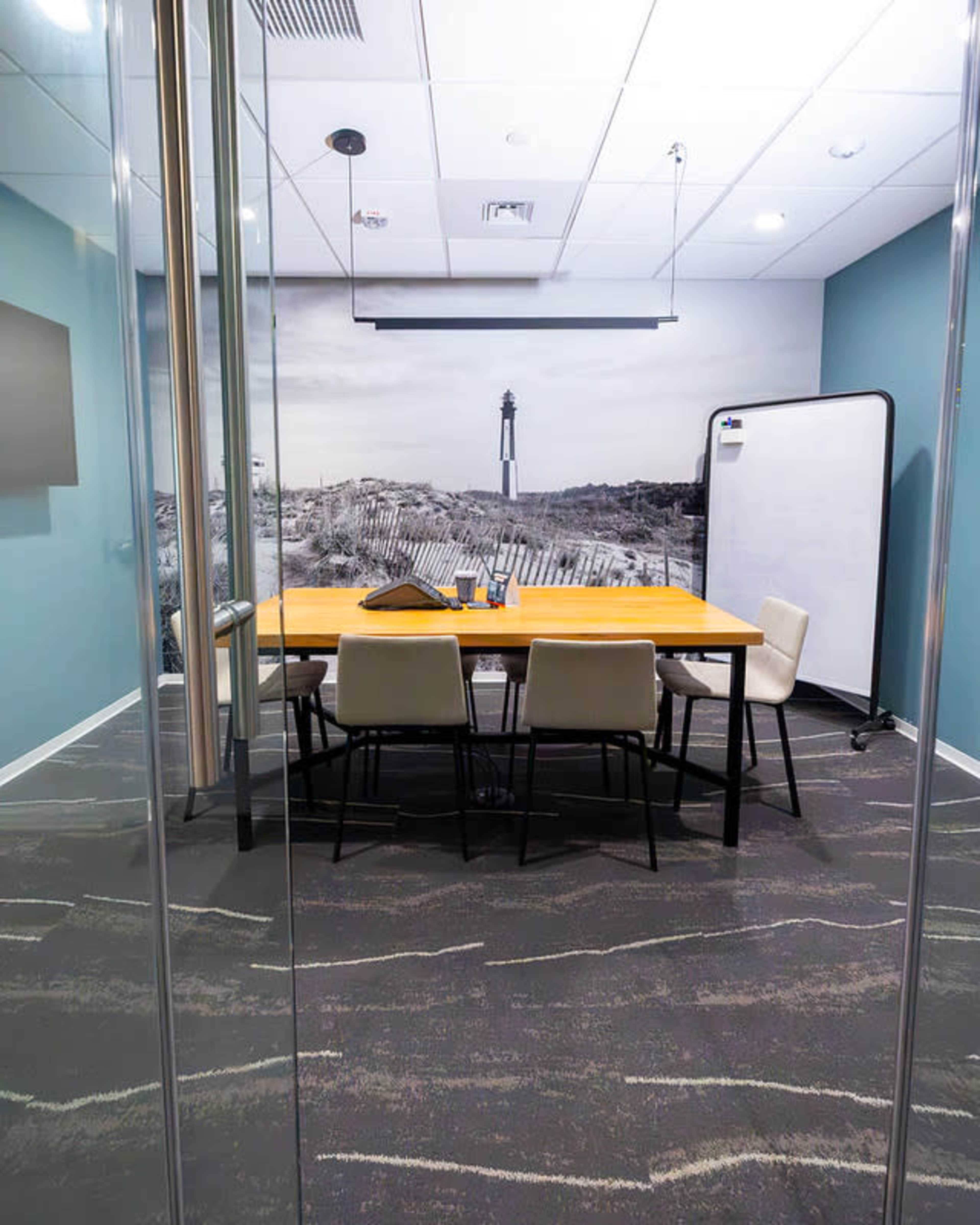 The image shows a modern conference room with a long wooden table surrounded by four chairs, a whiteboard, and a wall mural of a lighthouse and dunes in the background.